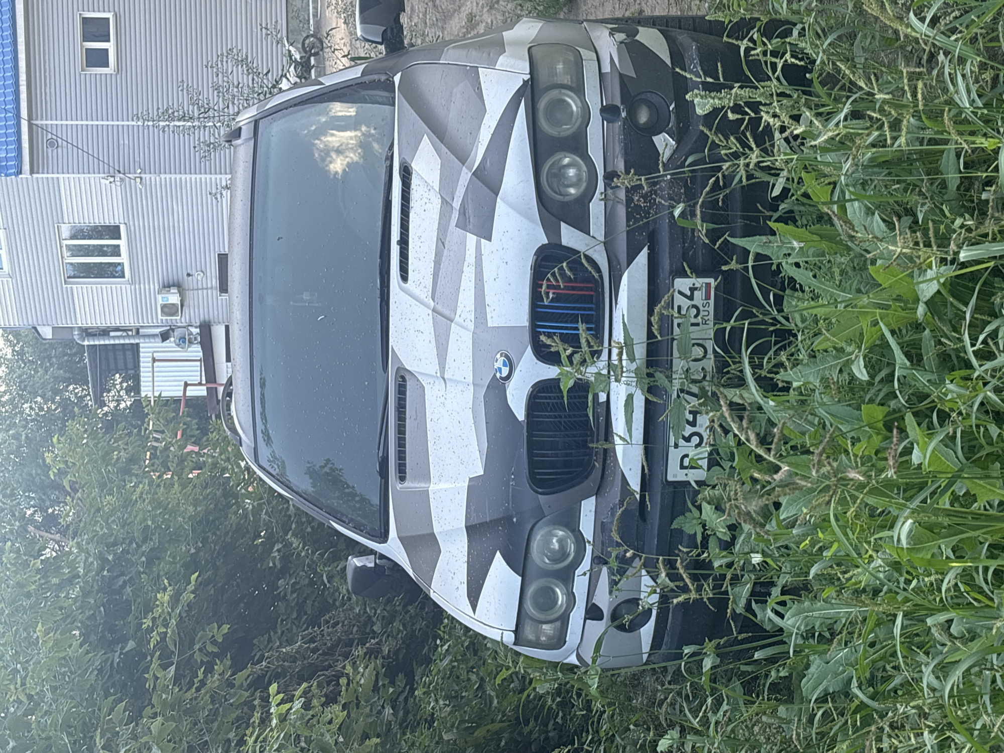 The image shows a heavily camouflaged BMW SUV parked in overgrown vegetation in the city of Ob, Russia. The vehicle is facing the camera, with its front end partially obscured by tall grass and weeds. The camouflage pattern on the car consists of geometric shapes in black, white, and grey. The front grille features the BMW logo and the M tri-color stripes (blue, red, and blue). The license plate, visible at the bottom center, reads "R347CO 154 RUS". In the background, a two-story building with light gray siding and a blue roof is visible. To the right of the building, a white dirt bike is parked. The overall environment suggests an outdoor, somewhat neglected setting, possibly a residential area or an unused lot.
