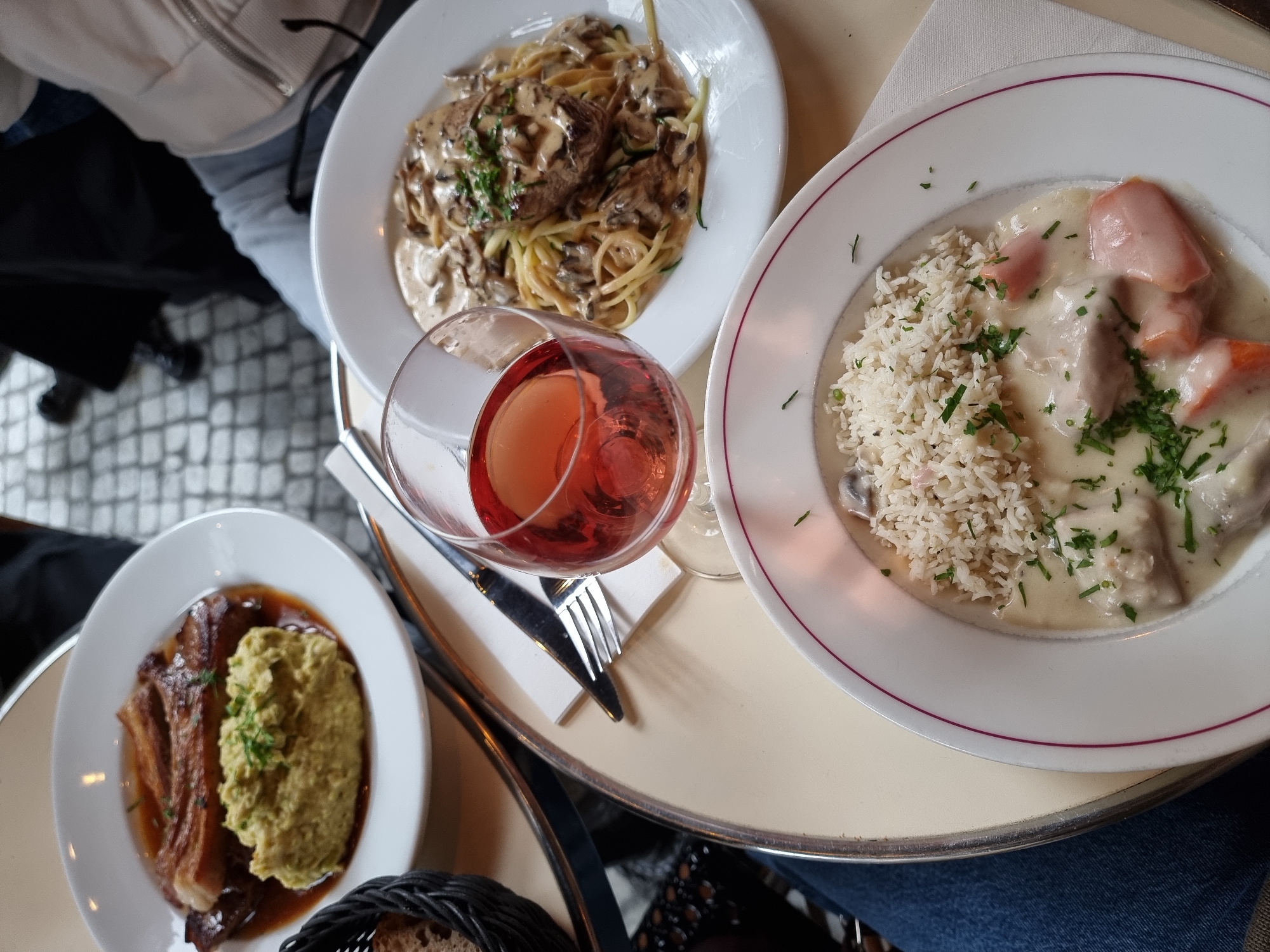The image depicts a table setting in a Parisian cafe. Three plates of food are visible, along with a glass of rosé wine, cutlery, and a bread basket. The food includes: pasta with a mushroom cream sauce, a creamy stew with rice, and a piece of meat with a side of green purée. The setting is likely outdoors, as suggested by the cobblestone pattern visible in the background. The presence of multiple plates and a wine glass indicates a dining experience, possibly a lunch or dinner.