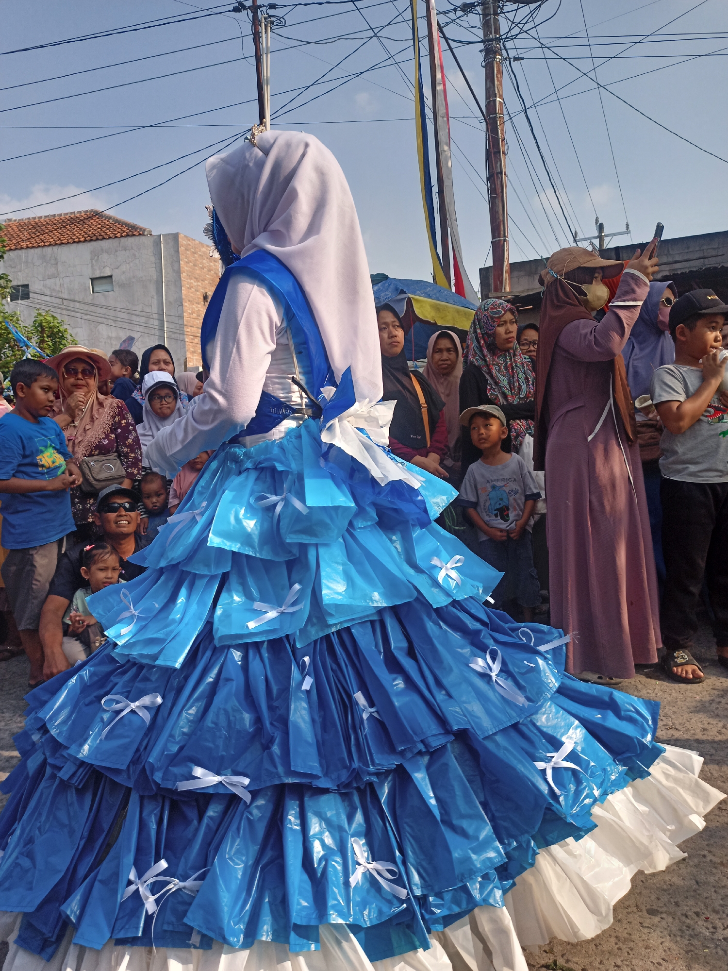 A person in a large, layered blue and white costume, likely for a festival, stands in the foreground. The costume resembles a full skirt with ruffles and small white bows. The person is wearing a light-colored hijab and a blue sash. In the background, a crowd of people, including children and adults, are gathered, some looking towards the person in costume. Some individuals are holding phones, possibly taking pictures or videos. The setting appears to be an outdoor street scene in a residential area, with buildings, a utility pole with wires, and flags visible. The sky is blue with some wispy clouds. The overall atmosphere suggests a community event or celebration. The location is identified as Saren, Indonesia.