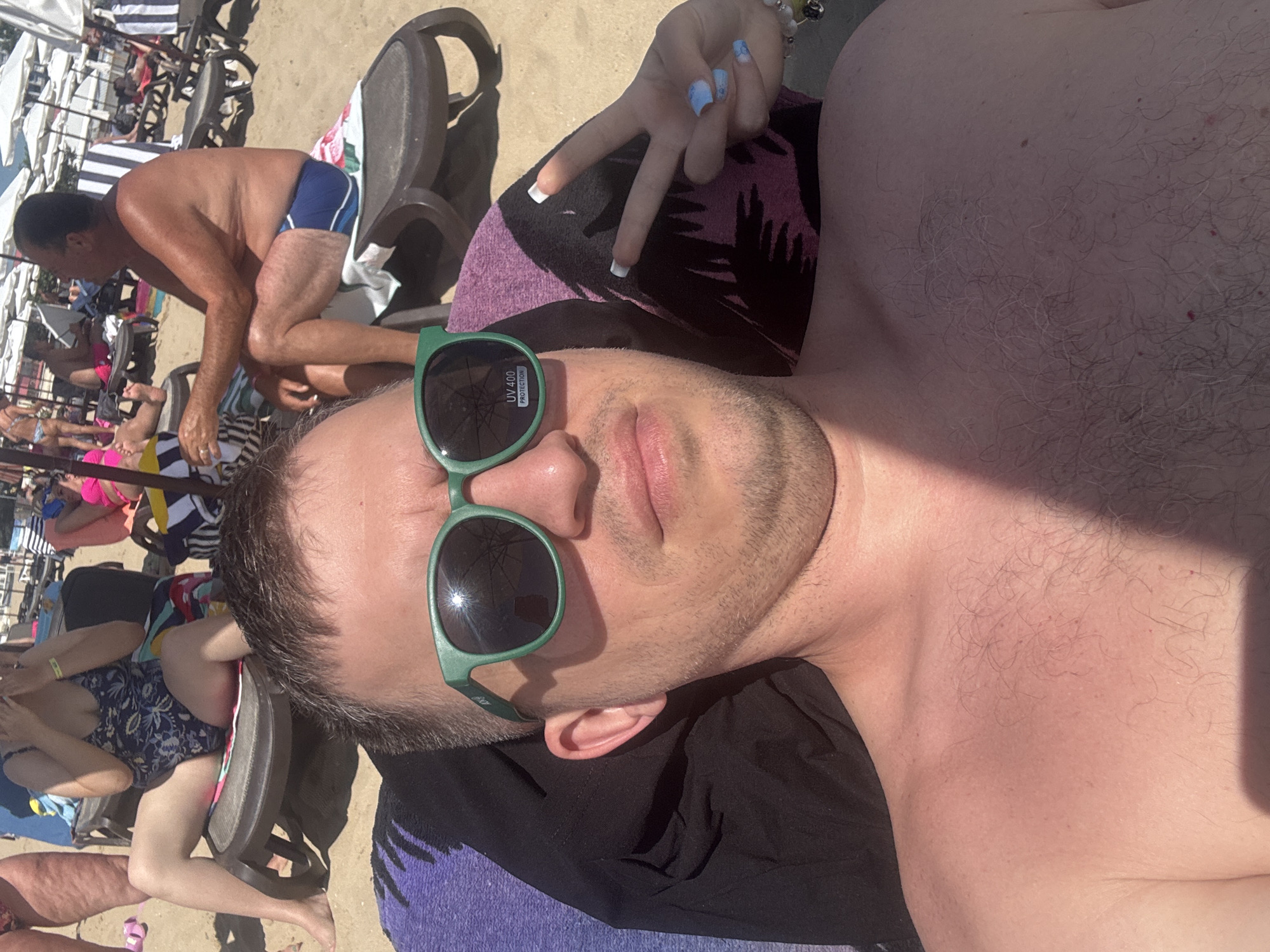 A man is lying on a beach towel on a lounge chair on the beach in Nesebar, Bulgaria. He is wearing green sunglasses and has his camera pointed towards himself, taking a selfie. In the background, other beachgoers are visible lounging on chairs or walking. The man's chest is visible, and a woman's hand with manicured nails is giving a peace sign near his head. The scene appears to be a sunny day, with beach umbrellas providing shade in the background.