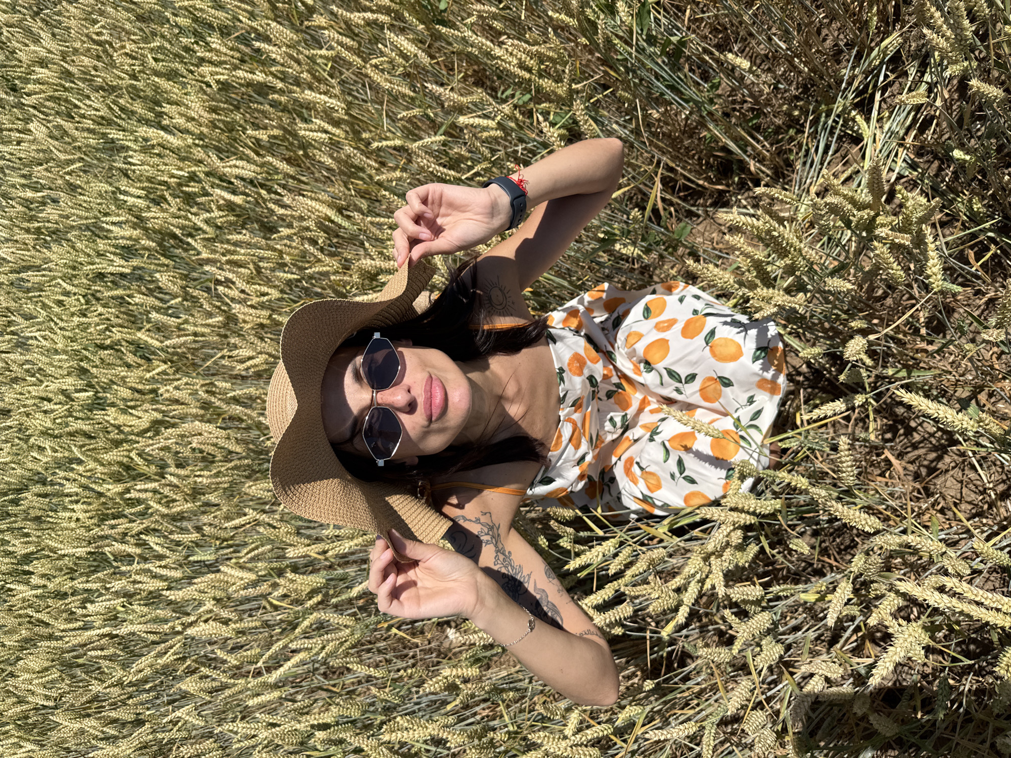 A woman wearing a straw hat and sunglasses sits in a field of wheat in Zgalevo, Bulgaria. She is wearing a white dress with an orange fruit pattern and is holding the brim of her hat. Her right arm is resting on her knee, and she has a tattoo on her left arm. The wheat stalks are tall and golden, and the sun is shining brightly.