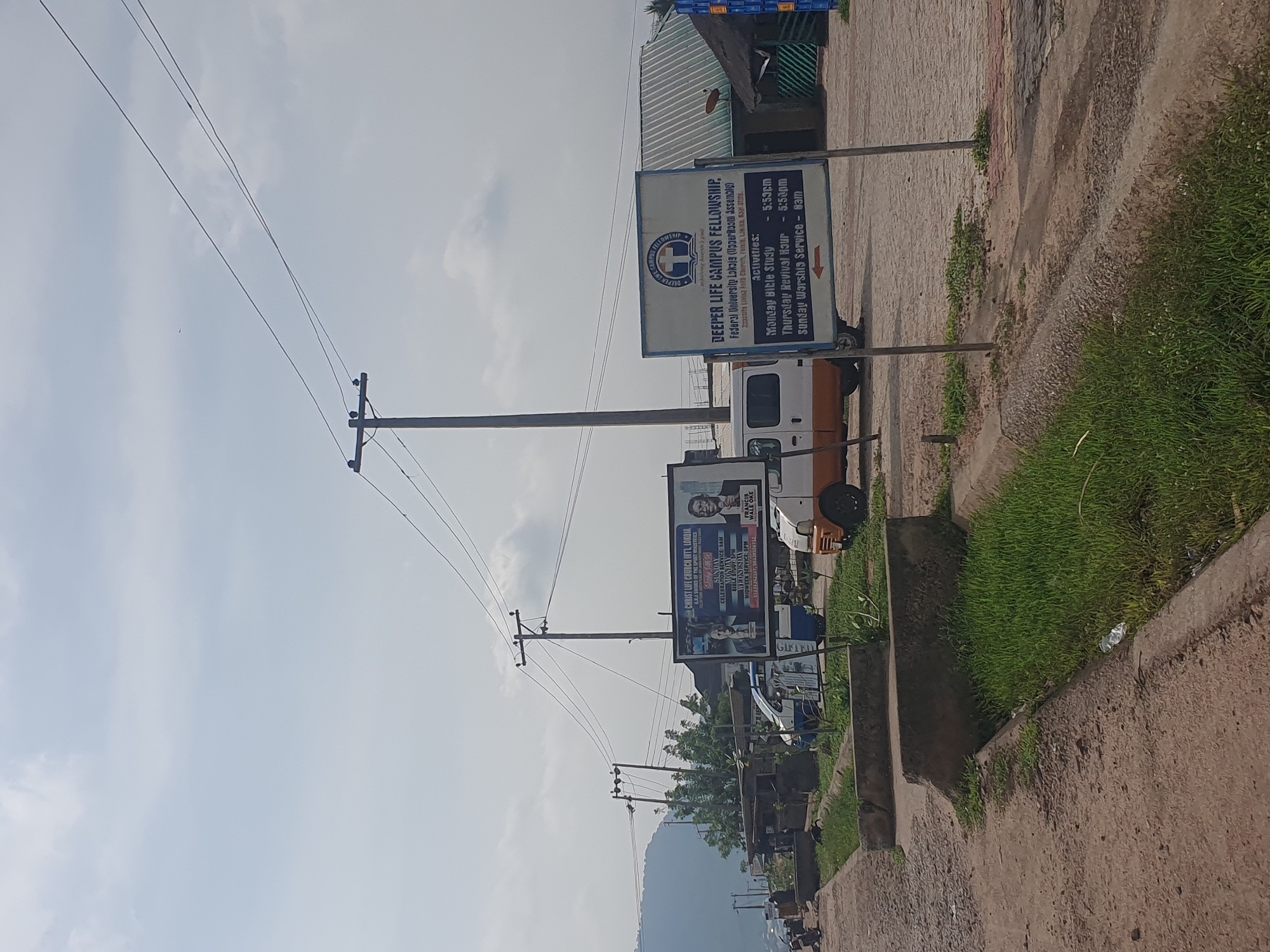 The image depicts a street scene in Lokoja, Nigeria, characterized by overcast skies and a mix of modern and older structures. In the foreground, a paved road or walkway is visible, bordered by a drainage ditch filled with lush green grass. Concrete barriers are strategically placed along the edge of the walkway. A prominent billboard stands on the right side of the image, displaying information about the "Deeper Life Campus Fellowship." The text on the billboard indicates the location as "Federal University Lokoja (Upper Room Assembly)" and lists activities with their corresponding times: "Monday Bible Study - 5:30pm," "Thursday Revival Hour - 5:30pm," and "Sunday Worship Service - 8am." Adjacent to the billboard, a green-roofed building with a thatched awning is visible.
Further down the street, a white and orange van is parked. To the left, a blue and white minivan or utility vehicle is also present. In the middle ground, there are several small structures, possibly shops or stalls, with corrugated metal roofing. Power lines crisscross the sky, supported by utility poles. A larger billboard with images of two men and text is situated to the left of the "Deeper Life Campus Fellowship" sign. In the background, a dense cluster of trees and vegetation can be seen, with a mountain or large hill rising in the far distance. A few individuals are sparsely scattered along the street, with some appearing to be walking or engaged in activity near the smaller structures. The overall impression is one of a functional, everyday urban environment in a Nigerian city.