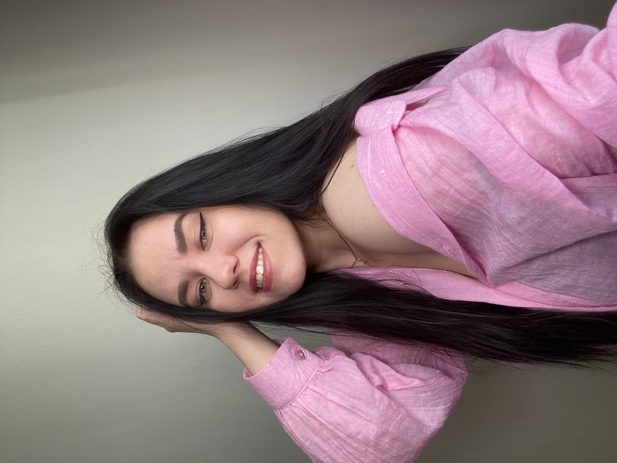 A young woman with long, straight black hair is smiling at the camera. She has fair skin, dark eyebrows, and light-colored eyes, wearing eyeliner. She is wearing a pink, button-down shirt or blouse that is loose-fitting and partially unbuttoned at the top. Her hand rests near her head, suggesting she might be lying down or leaning. The background is a plain, light-colored wall. There is no information to confirm that the location is in the city of Chaikovsky, Russia.