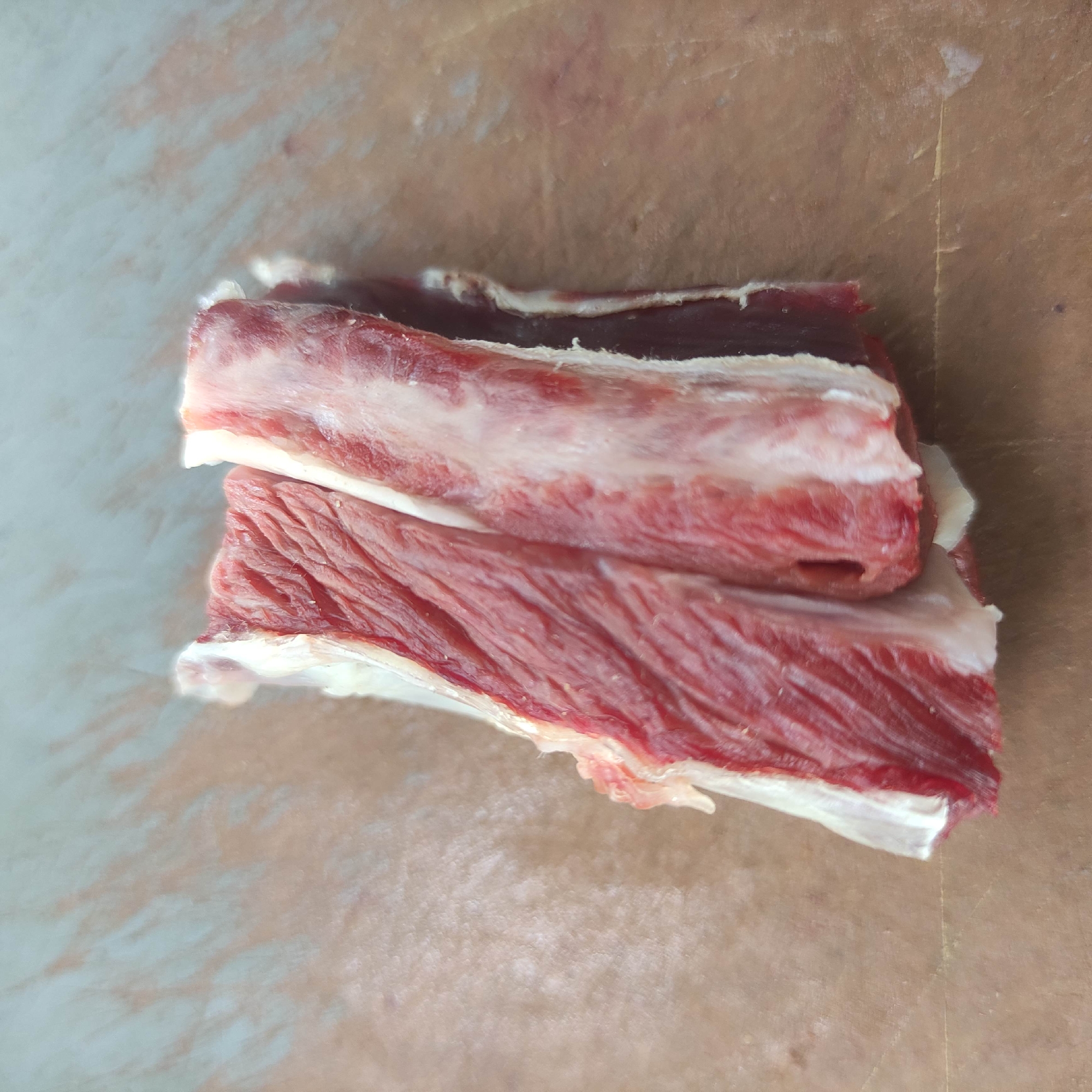 A close-up, overhead shot shows two pieces of raw, fatty meat with visible muscle fibers. The meat is placed on a textured, light brown surface that appears to be parchment paper or butcher paper. The meat is deep red in color with thick, white streaks of fat running through it. The lighting is bright and even, creating subtle shadows and highlighting the texture of the meat and the surface. The image is focused on the meat, with the background slightly out of focus. There are no people or any other objects visible in the frame. The location is not identifiable from the image.
