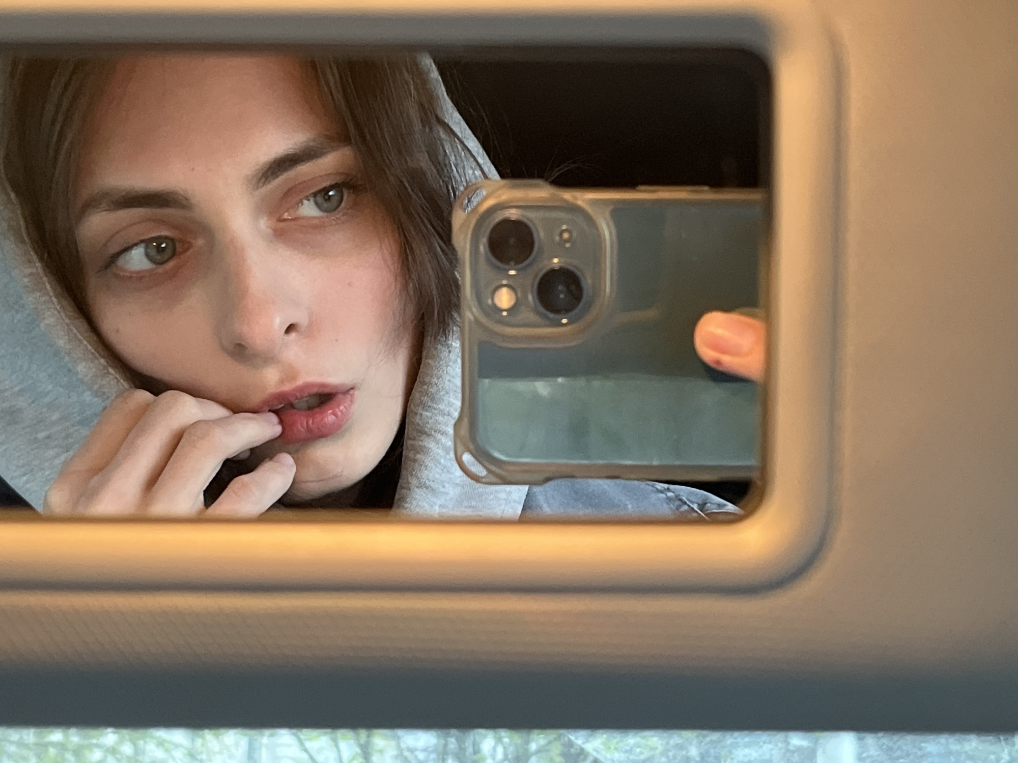 A woman with long dark hair wearing a grey hooded sweatshirt is taking a selfie. Her fingers are near her lips. The selfie is taken in a mirror, with a smartphone visible in the reflection. The mirror appears to be a vanity mirror inside a vehicle. It is not possible to determine if the photo was taken in Krasnoyarsk, Russia.
