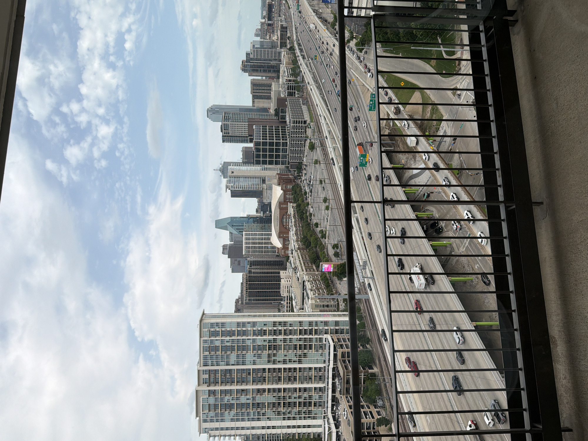 From a high vantage point, likely a balcony, a cityscape view of Dallas, Texas, is presented. Skyscrapers and office buildings form the skyline, appearing under a partially cloudy sky. Below, a large, multi-lane highway is visible, filled with moving vehicles. The perspective is somewhat rotated, with the balcony railing being the foreground element, partially obscuring the lower right of the scene.