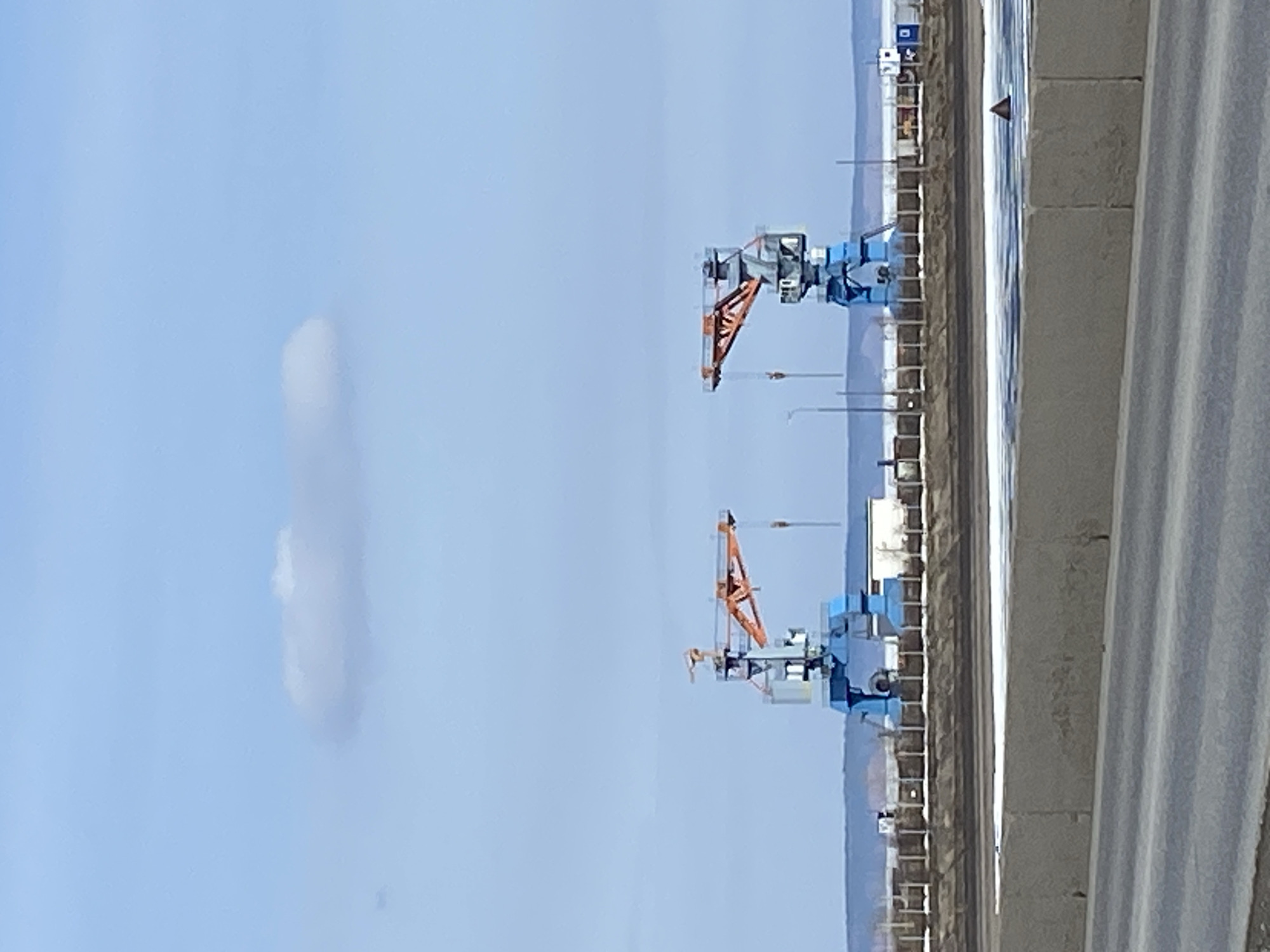 The image shows two rail-mounted gantry cranes in what appears to be an industrial area. The cranes are painted blue and have orange booms. In the background, there are other industrial buildings and structures. A cloud is visible in the blue sky. A concrete barrier is in the foreground. It is not possible to confirm the location of Chaikovsky, Russia based on the provided image alone.