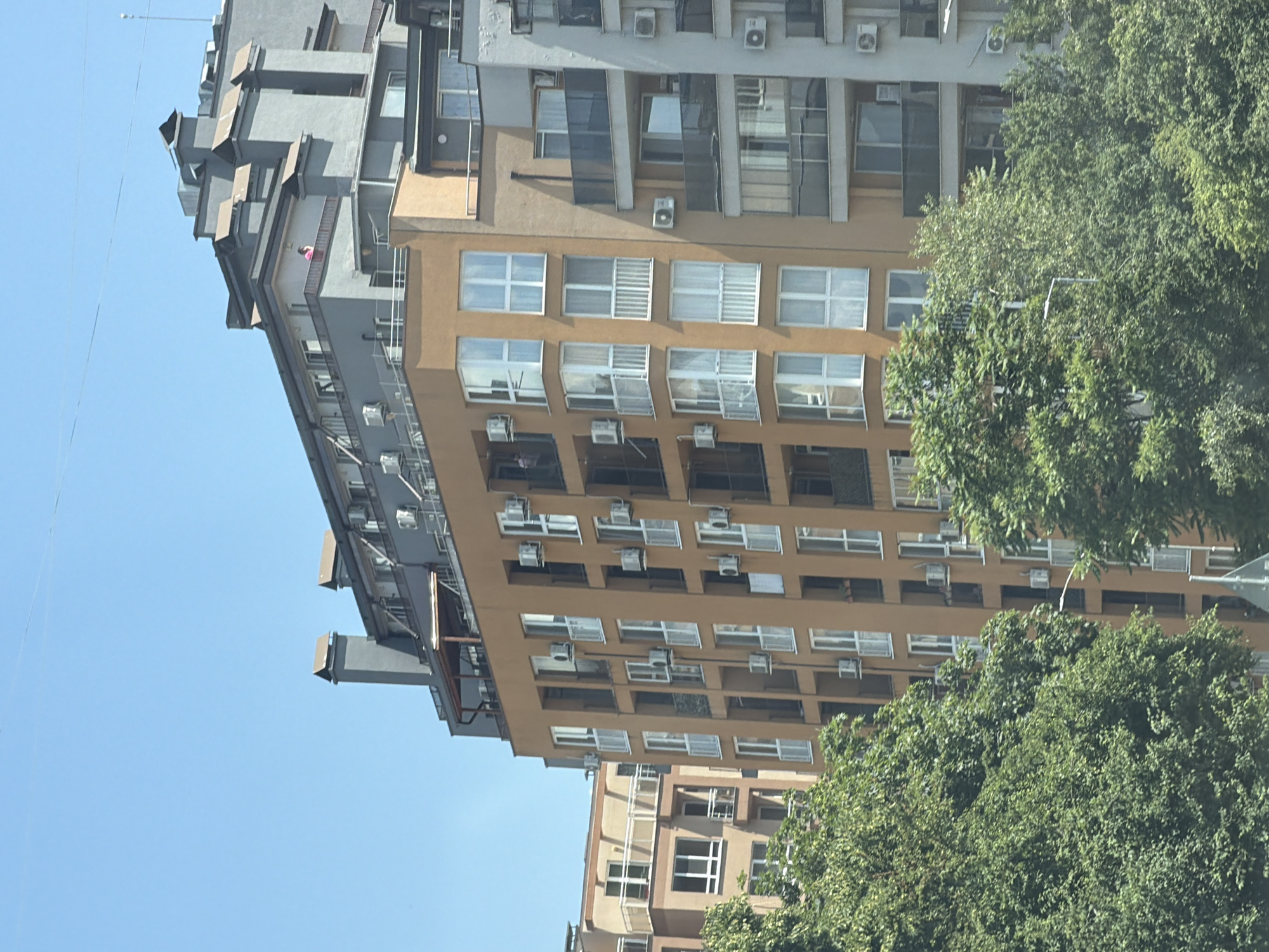 A tall, modern apartment building with an orange facade is the primary subject, seen from a slightly low angle, partially obscured by lush green trees in the foreground. The building features numerous balconies with white railings and many windows, some with white curtains. Multiple air conditioning units are visible attached to the exterior walls. Several other buildings, one with a lighter colored facade and balconies with glass railings, and another grey building with a more complex, stepped roofline and chimneys, are partially visible to the left and right, respectively. A person wearing a pink top can be seen standing on a balcony on the grey building. The sky above is a clear, bright blue. The scene appears to be a residential area in Sofia, Bulgaria, on a sunny day.