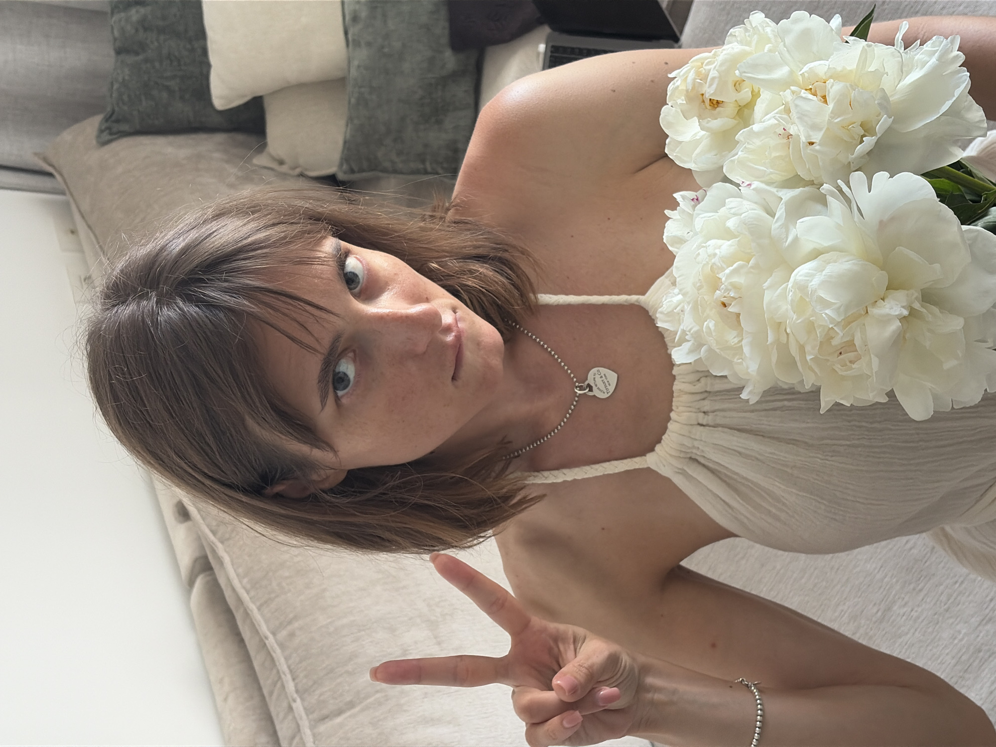 A young woman with brown hair and blue eyes is shown from the chest up, holding a bouquet of white peonies and making a peace sign with her left hand. She is wearing a white halter-neck dress and a silver necklace with a heart-shaped pendant. The background includes a couch with several pillows. The image appears to be taken indoors, possibly in a residential setting. There is no visible indication of the location being Tel-Aviv, Israel.