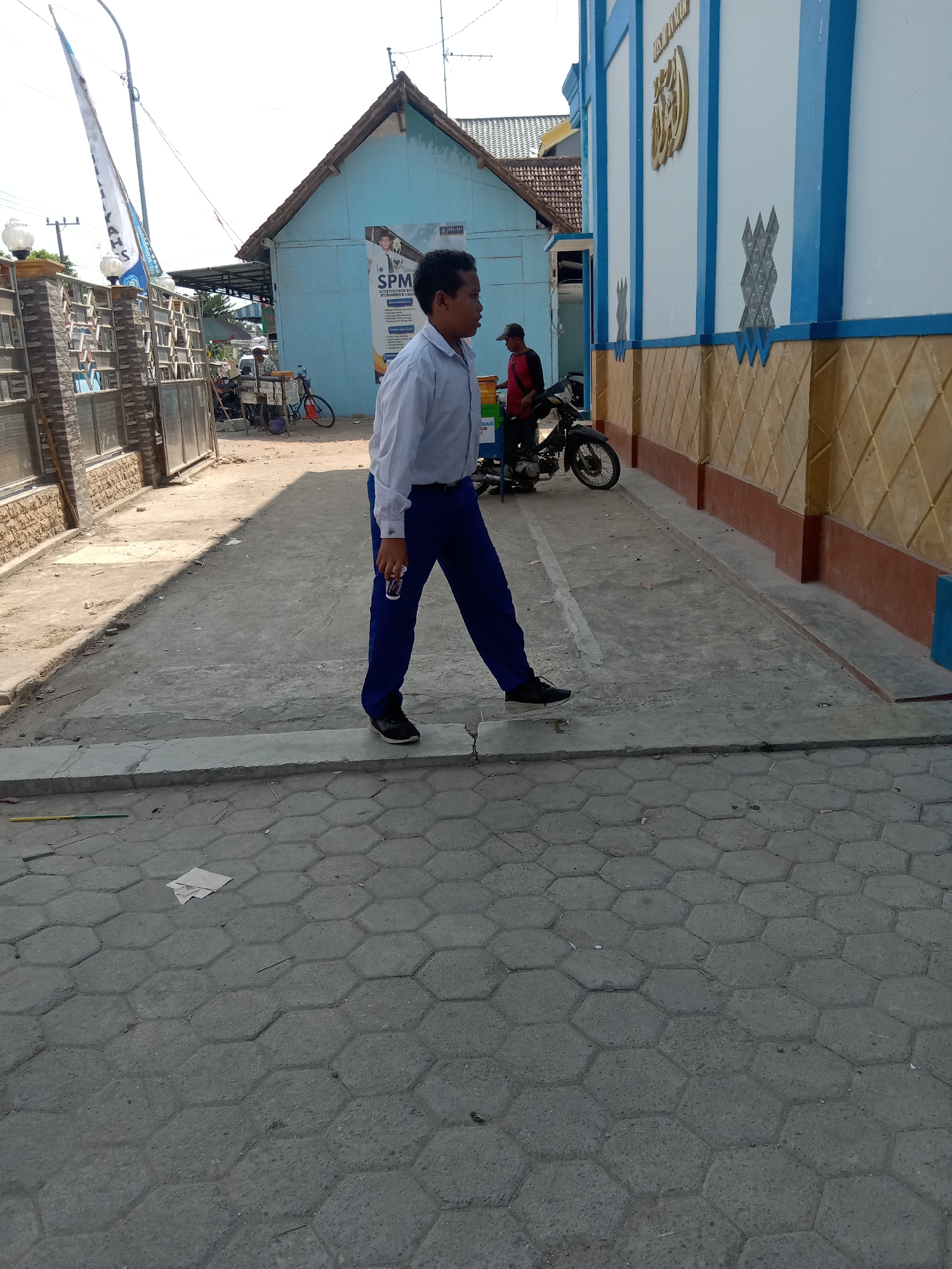 In Sugihwaras, Indonesia, a man in a white collared shirt and blue pants walks across a paved area. He is holding a white cup. Behind him, a person wearing a red shirt and a cap stands next to a motorcycle. A light blue building with a brown and yellow patterned base is visible to the right, and a similar blue building with a brown roof is to the left, featuring a poster on its side. A metal fence with stone pillars stands to the far left, and two flags are attached to poles in front of it. The ground is paved with hexagonal concrete blocks.