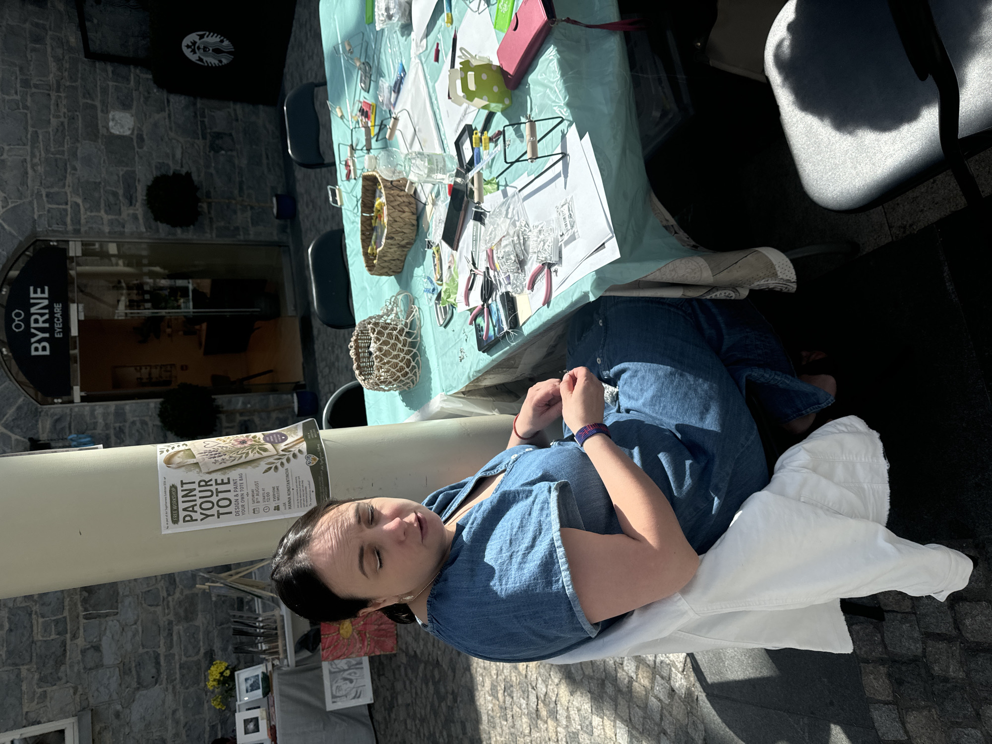 In the city of Kilkenny, Ireland, a woman with dark hair is seated at a table covered with a teal tablecloth. She is wearing a blue denim dress and a white jacket is draped over her chair. The table is laden with various craft supplies, including pliers, beads, papers, and baskets. A sign on a pillar behind her advertises a "Paint Your Tote" free workshop. In the background, a building with "BYRNE EYECARE" signage is visible. The scene appears to be outdoors, with sunlight casting shadows on the ground.