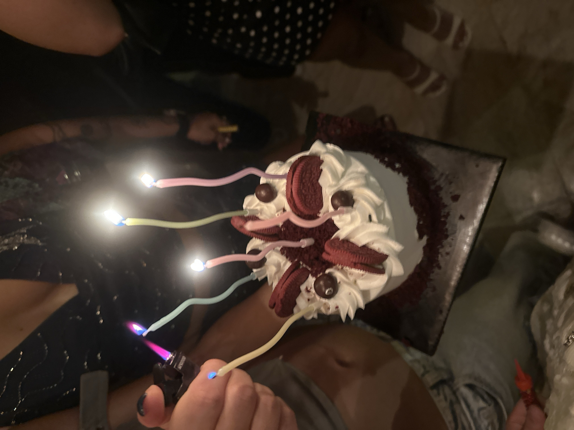 The image depicts a red velvet cake decorated with whipped cream, Oreo cookies, and chocolate balls, adorned with lit birthday candles. A hand is holding a lighter to light the candles. The scene appears to be a birthday celebration. Location: North Kuta, Indonesia. Additional people, wearing various casual outfits, are visible around the cake. The background is somewhat blurred, suggesting an indoor setting with warm lighting.