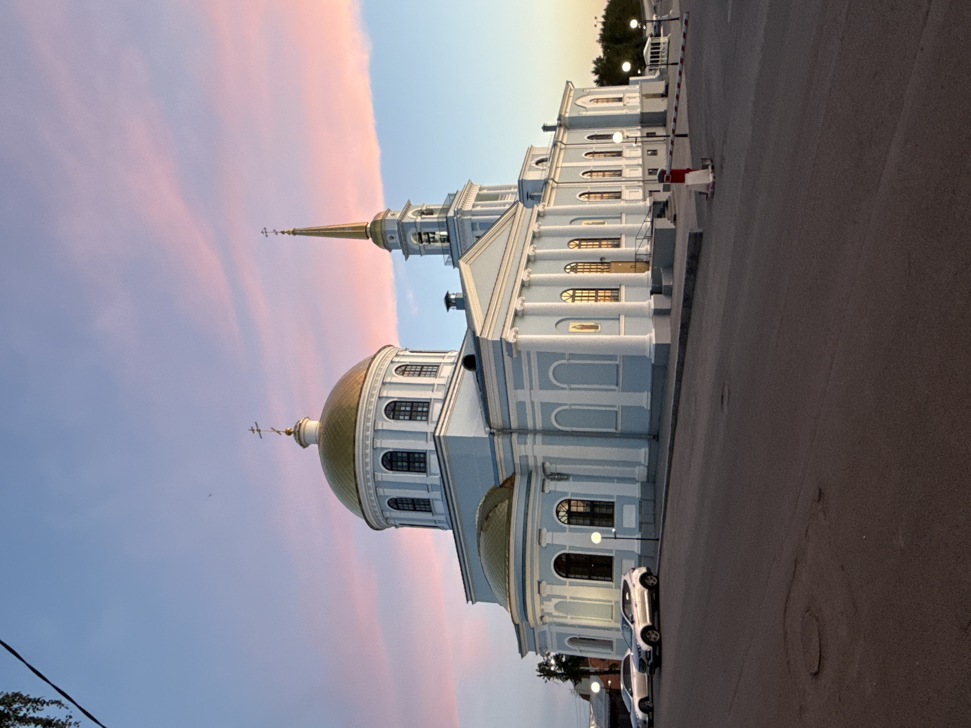 The image captures a grand Orthodox church, likely situated in Votkinsk, Russia, bathed in the soft light of either dawn or dusk. The main subject is a large, stately building painted in light blue and white, adorned with classical architectural features such as prominent columns, arched windows, and intricate cornices. It is characterized by multiple gleaming golden domes, including a large central dome and two smaller ones, each topped with an Orthodox cross. A tall, slender spire with another cross rises from a bell tower towards the right side of the church structure.
The sky presents a beautiful gradient, transitioning from soft pink and orange clouds on the left, indicative of the sun's low position, to a clearer, lighter blue on the right. The scene is illuminated by glowing streetlights and light spilling from the church windows, creating a warm and serene atmosphere against the twilight sky.
Adjacent to the church, an asphalt road or parking lot extends, where several white cars are visibly parked. A red and white striped barrier can be seen further down the road, suggesting a controlled entry point. The overall setting is tranquil, with no people visible, allowing the architectural splendor of the church to be the central focus under the dramatic sky. There is no visible text in the image.