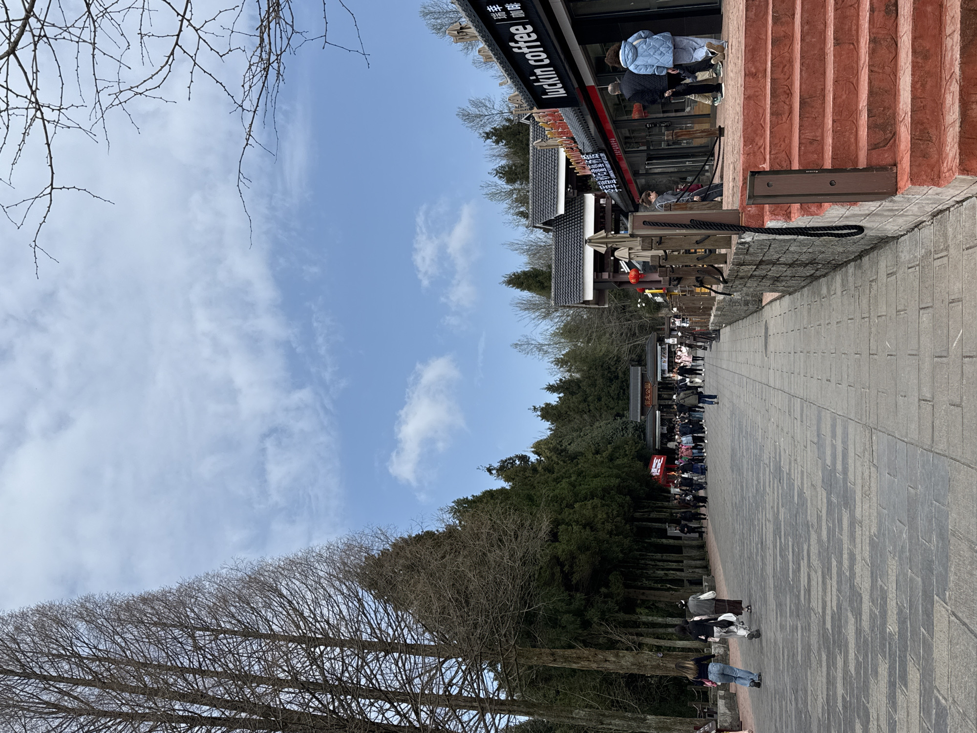 The image depicts an outdoor pedestrian area in Wulingyuan, China, under a partially cloudy blue sky. A wide pathway of rectangular grey flagstones extends into the distance, flanked by rows of tall trees. On the left, several large deciduous trees with bare branches line the path. To the right, a series of commercial buildings with dark, traditional Chinese-style tiled roofs are present. One building features a black facade with red trim and displays a prominent "luckin coffee" sign in white English and Chinese characters. In front of these buildings, a raised platform with five red-painted concrete steps is visible, alongside a low stone wall with dark wooden posts and a thick black rope. Red lanterns hang from a nearby structure.
Numerous individuals, primarily adults, are distributed across the scene. In the foreground on the left, three individuals are walking away from the viewer. Near the "luckin coffee" establishment on the right, three individuals are standing or walking towards the building, one wearing a light blue puffer jacket with their back to the camera. Further down the paved path, a larger group of approximately 20 people is gathered near a traditional Chinese-style gate structure, possibly an entrance to a park, where a red banner is also visible. The overall scene suggests a busy tourist or public access area.