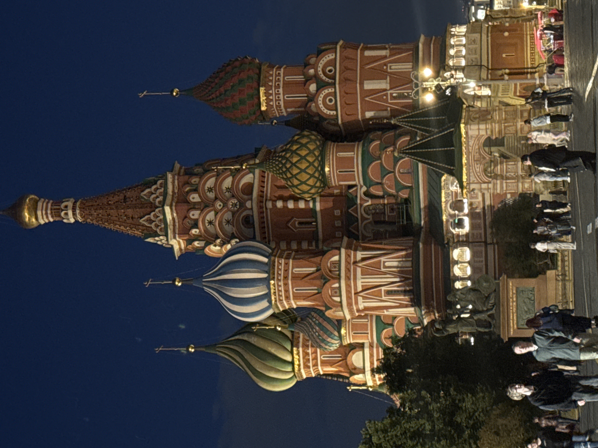This is a nighttime photograph of Saint Basil's Cathedral in Red Square, Moscow, Russia. The cathedral, a UNESCO World Heritage site, is illuminated, showcasing its distinctive onion domes, vibrant colors, and intricate architectural details. The sky above is a deep blue, indicative of twilight or night.
In the foreground, several people are walking on a cobblestone street. Some are looking towards the cathedral, while others appear to be engaged in conversation or walking purposefully. The lighting suggests a public space with pedestrian activity. There is also a statue in the middle ground, possibly a monument. Trees are visible on the left side of the image, partially obscuring the cathedral. Streetlights and lights from within the cathedral add to the ambiance. In the far right, there are small market stalls or food vendors, also illuminated. The overall impression is of a well-known landmark bustling with evening activity.