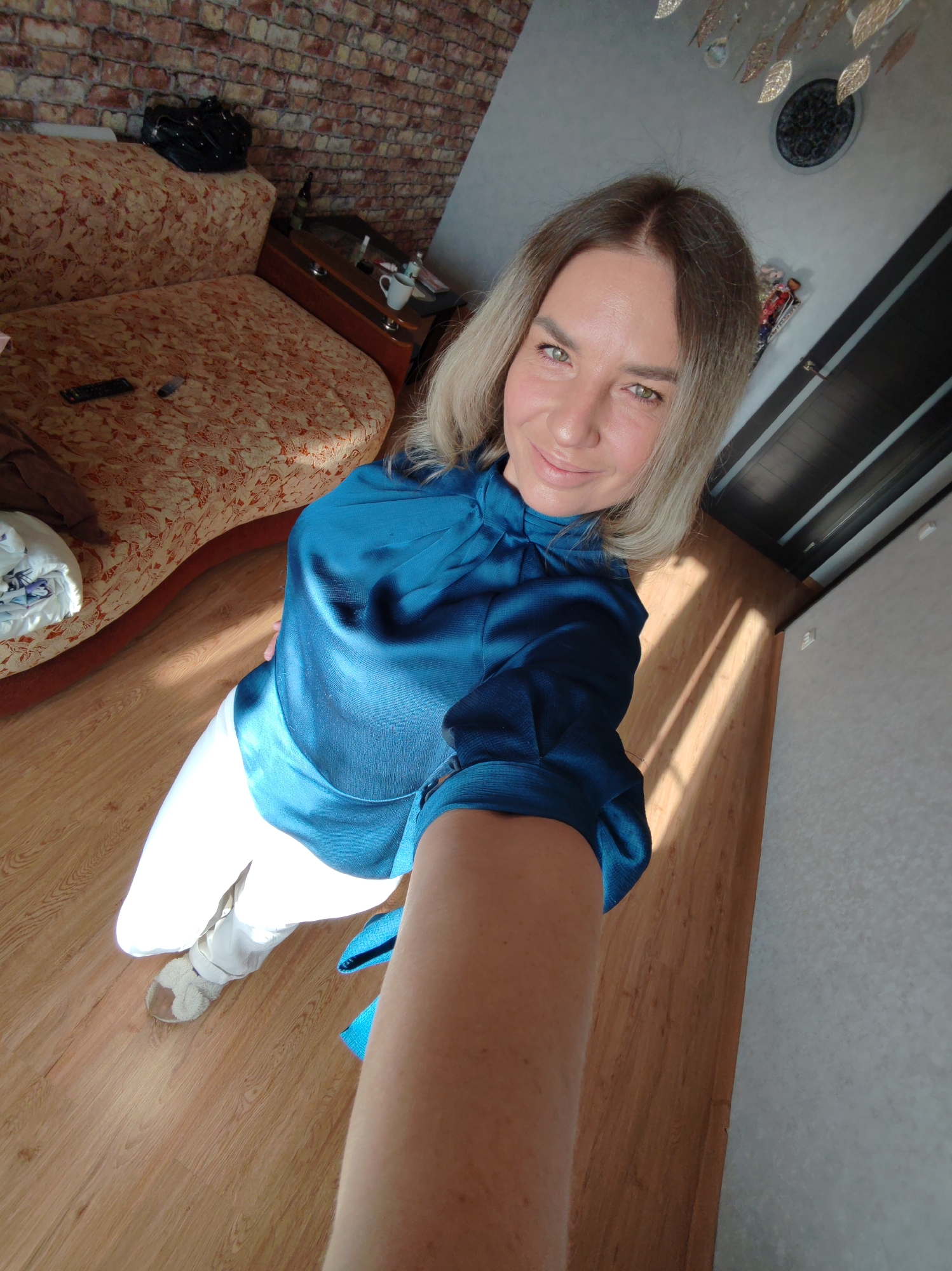 A woman with long, ash blonde hair and bright green eyes is captured in a high-angle selfie, smiling gently at the camera. She is wearing a teal blue long-sleeved satin blouse with a high neck and white pants. On her feet, she has light-colored, possibly fluffy, slippers.
The setting appears to be an indoor residential space, likely a living room or bedroom in Petropavlovsk-Kamchatsky, Russia. The background features a couch with an orange and white patterned upholstery, upon which a TV remote control is visible. Behind the couch, a wall with brick-patterned wallpaper adds texture to the room. To the right of the couch, a small wooden side table with a glass top holds a white mug, a dark bottle, and other small items.
Above the woman's head, a decorative chandelier with gold leaf-like elements hangs from the ceiling, and a dark, circular wall decoration is also visible. The floor is covered in light-colored wooden laminate, across which strong diagonal sunbeams are cast, indicating it is daytime. A dark wooden door with horizontal panels is partially visible on the far right. The woman is holding the camera herself, creating a self-portrait. There is no visible text in the image.