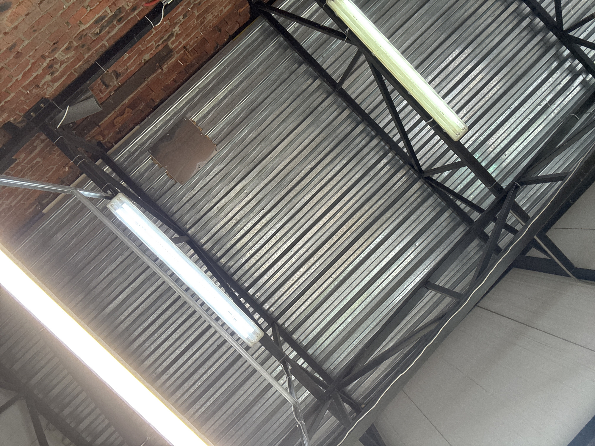 The image presents an upward, diagonal view of an interior ceiling structure, characteristic of an industrial, workshop, or warehouse setting. The dominant feature is a corrugated metal ceiling, composed of galvanized steel panels running diagonally across the frame. These panels are supported by a framework of black painted metal trusses and beams, which crisscross the ceiling, creating a sturdy yet exposed structural aesthetic.
In the upper left portion of the image, a section of an exposed brick wall is visible. Black electrical conduits and wiring are installed along this brick wall and also run across some of the metal beams, indicating functional infrastructure. A small, rectangular, dark metal plate is affixed flush with the corrugated ceiling in the upper-central area.
Three fluorescent tube light fixtures are prominently featured. One, intensely bright, is partially visible in the lower-left, while two other fully visible fixtures are positioned centrally and towards the upper-right. All the lights appear to be active, illuminating the metallic ceiling surface.
No people or specific activities are visible in the frame, indicating a focus purely on the architectural and structural elements of the space. The materials and construction suggest a practical, robust environment. No discernible text is present in the image. The scene depicts an interior space, located in Chaikovsky, Russia.