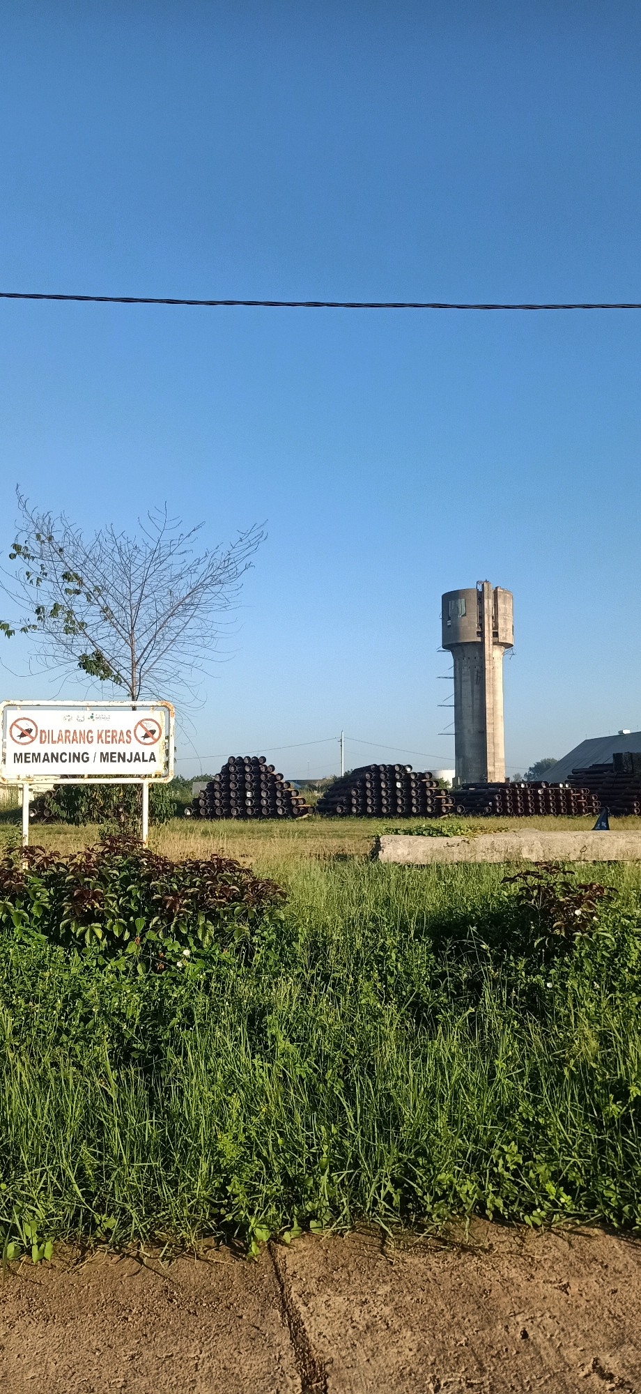 The image depicts an outdoor scene in Keudekruenggeukueh, Indonesia, under a clear blue sky. In the foreground, tall green grass and some leafy plants are visible, bordering a sandy or dirt pathway. To the left, a white sign with red lettering and symbols is posted on a pole. The sign has two circular symbols with a diagonal line through them, indicating prohibition. The text on the sign reads "DILARANG KERAS MEMANCING / MENJALA". Behind the sign and grass, a bare-branched tree stands.
Further in the background, several large stacks of dark, cylindrical objects, likely pipes, are arranged on the ground. To the right, a tall, concrete water tower with a distinct architectural style is prominent. In the far background, a building with a dark roof is visible. Overhead, a dark wire stretches across the frame. The overall environment suggests an industrial or utility area adjacent to a natural landscape. There are no people or animals clearly visible in the image.