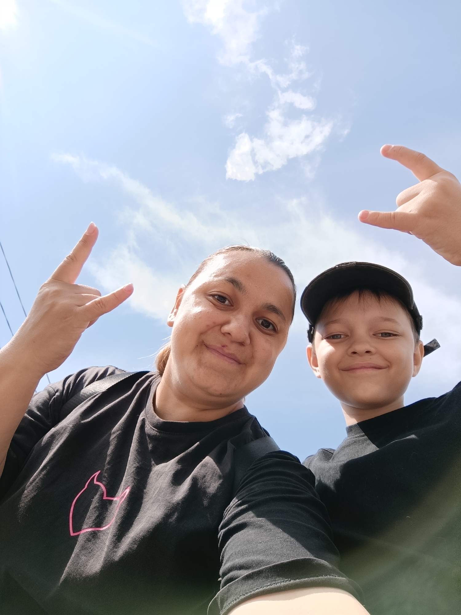 A low-angle, close-up selfie shows a woman and a boy against a bright blue sky with scattered clouds. Both are wearing black t-shirts and making a "rock on" hand gesture. The woman, on the left, has her hair pulled back and is smiling at the camera. The boy, on the right, is wearing a black baseball cap and grinning. The background suggests an outdoor setting, possibly in Chaikovsky, Russia, given the context, though no specific landmarks are visible. The lighting indicates it is daytime, with the sun creating lens flare in the upper left corner.