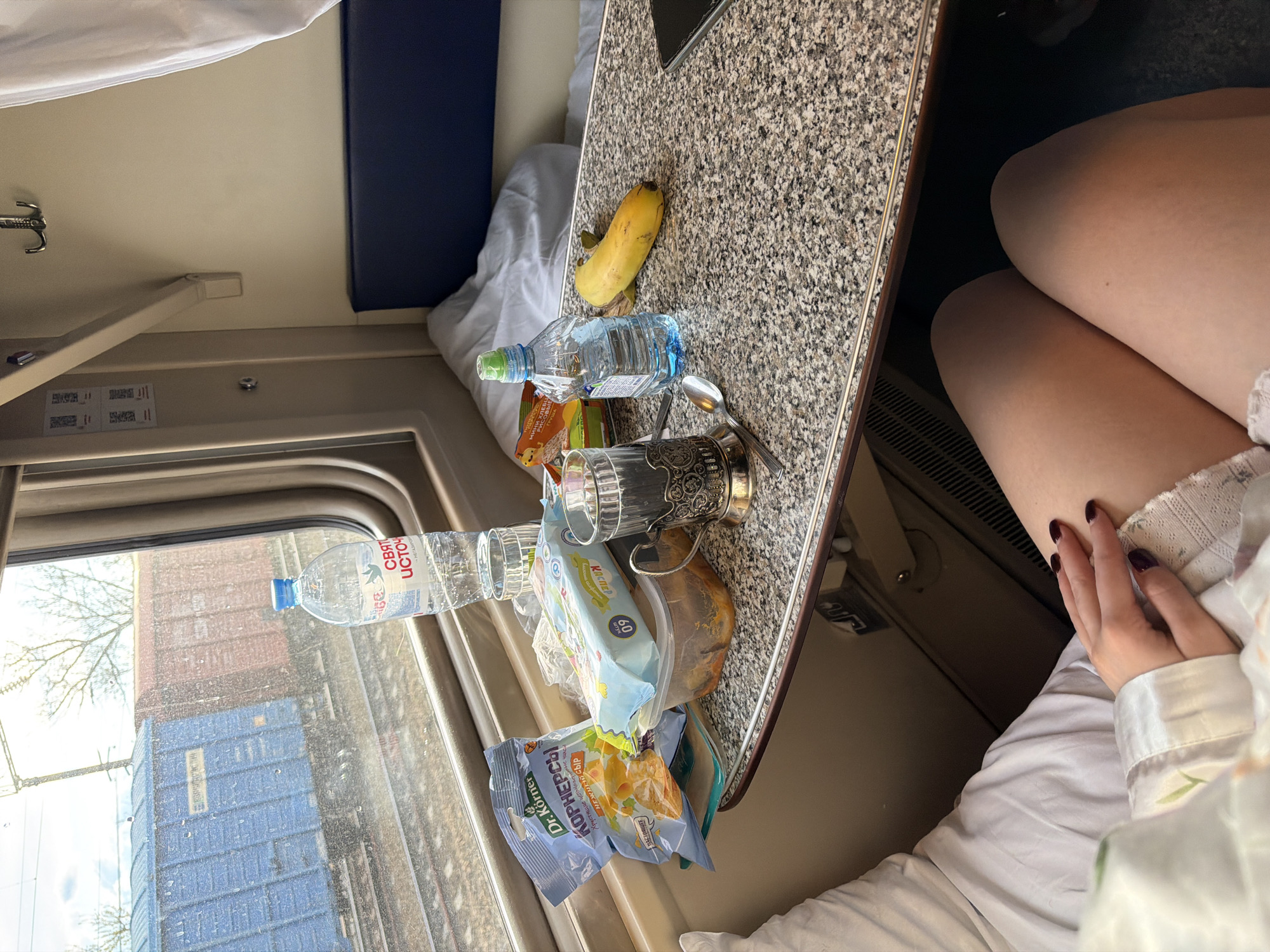 The scene is an interior view of a train compartment, likely taken from the perspective of a seated passenger. A table, covered in a speckled countertop, dominates the foreground and is laden with various items: a banana, a bottle of water, several packages of chips and snacks, a drinking glass in a metal holder with a spoon. The window to the left reveals a view of train tracks with cargo containers. A second bottle of water is visible through the window. Part of a blue cushioned seat can be seen in the background. A woman's thighs are also visible, suggesting she's seated near the table with one hand resting on her leg. The presence of these items suggests the passenger is on a journey.
I am unable to ascertain the exact location (Krasnyy Sulin, Russia) from the visual elements within the image. I am also unable to perform geolocation analysis using the image.