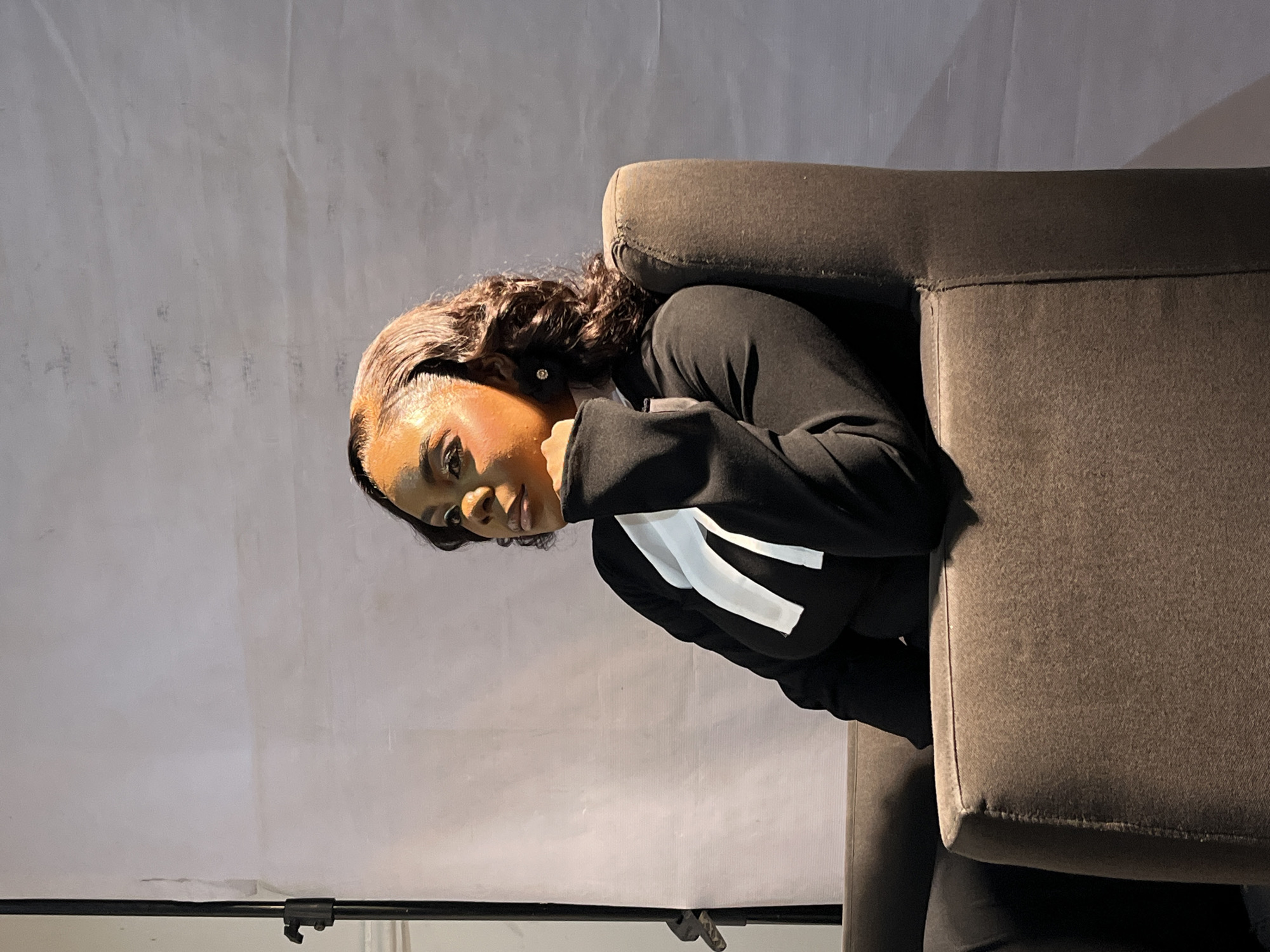 A medium shot shows a woman sitting in a dark brown armchair against a light gray backdrop. The woman is looking to her left with her chin resting on her hand. She has dark curly hair and is wearing a black jacket with a white V-shaped collar. She has on dramatic eye makeup and subtle lipstick. In the background, a black photography stand is visible on the left side of the frame. The lighting casts shadows on the backdrop and highlights the woman's face and hair.
