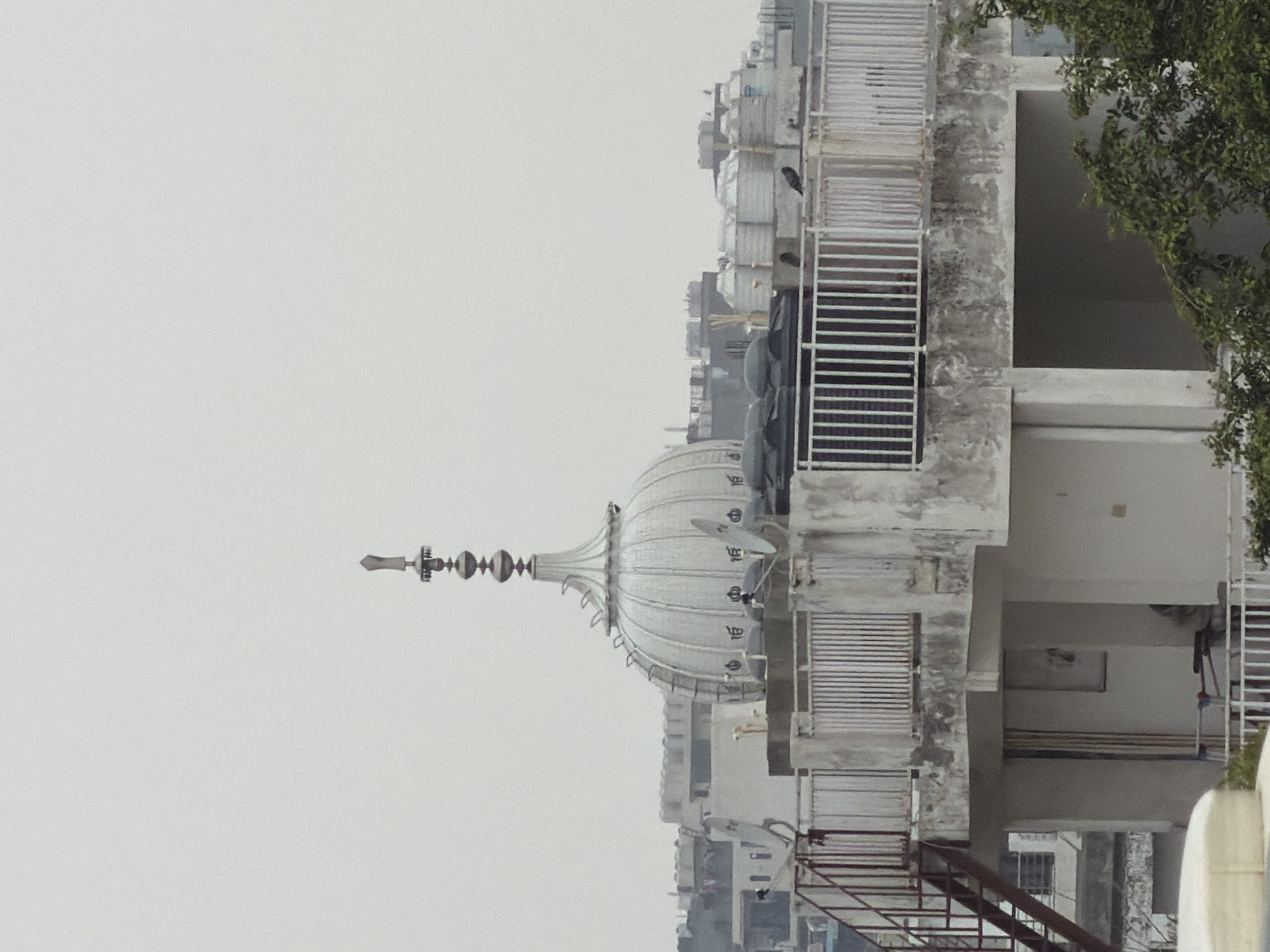 A telephoto shot captures a multi-tiered building structure topped with water tanks and a prominent, ornate dome. The dome, a central focal point, is silver-colored and intricately detailed with ridges and decorative elements, culminating in a sharp finial. Surrounding the dome are several satellite dishes and a white railing encircling the rooftop. In the background, a hazy cityscape of Delhi, India, is visible, with other buildings and structures suggesting an urban environment. The sky is overcast and pale, casting a muted light on the scene. A metal staircase is attached to the side of the building.