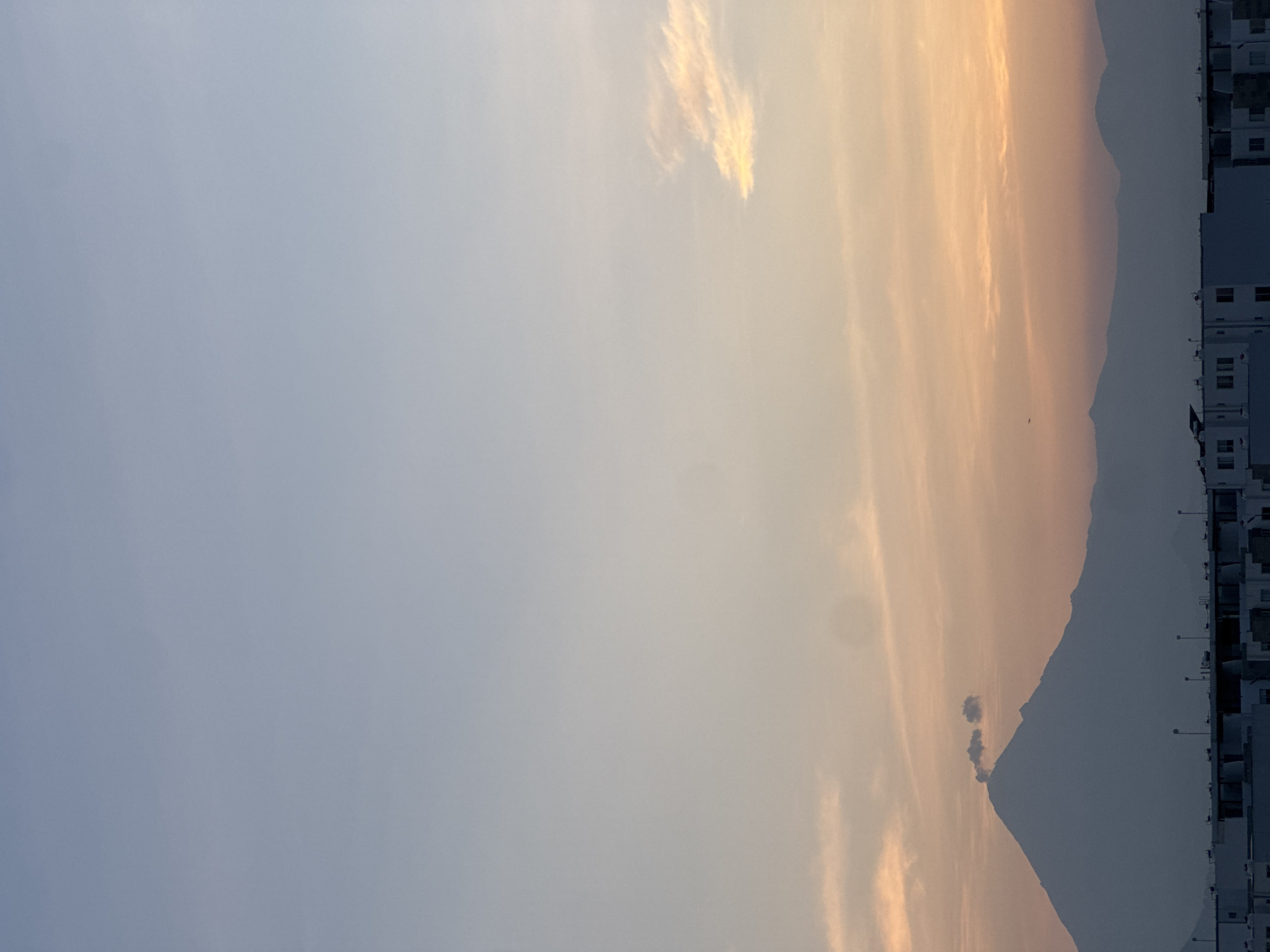 The image depicts a landscape at sunset or sunrise. In the background, there is a mountain range. To the right of the mountains, there are buildings, possibly residential structures. The sky is a gradient of colors, ranging from light blue to orange. There are some clouds in the sky. Based on the location provided, San Francisco Ocotlán, Mexico, the mountain range is likely part of the landscape in that region.