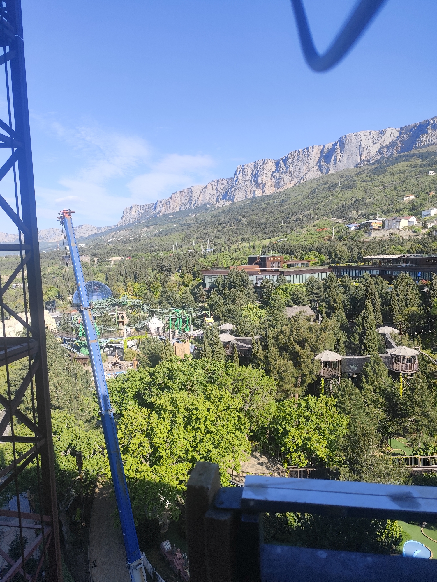 The image depicts a view of the landscape near Симеизский поселковый совет, Ukraine. In the background are mountains against a clear blue sky. Closer to the foreground, a large hotel complex is situated amidst dense trees. A portion of a green roller coaster, a geodesic dome, and several tree houses can also be seen. A blue crane is visible on the left side of the frame. The photograph appears to be taken from a high vantage point, possibly a tower or tall building, with a partial view of the structure's framework visible in the foreground.