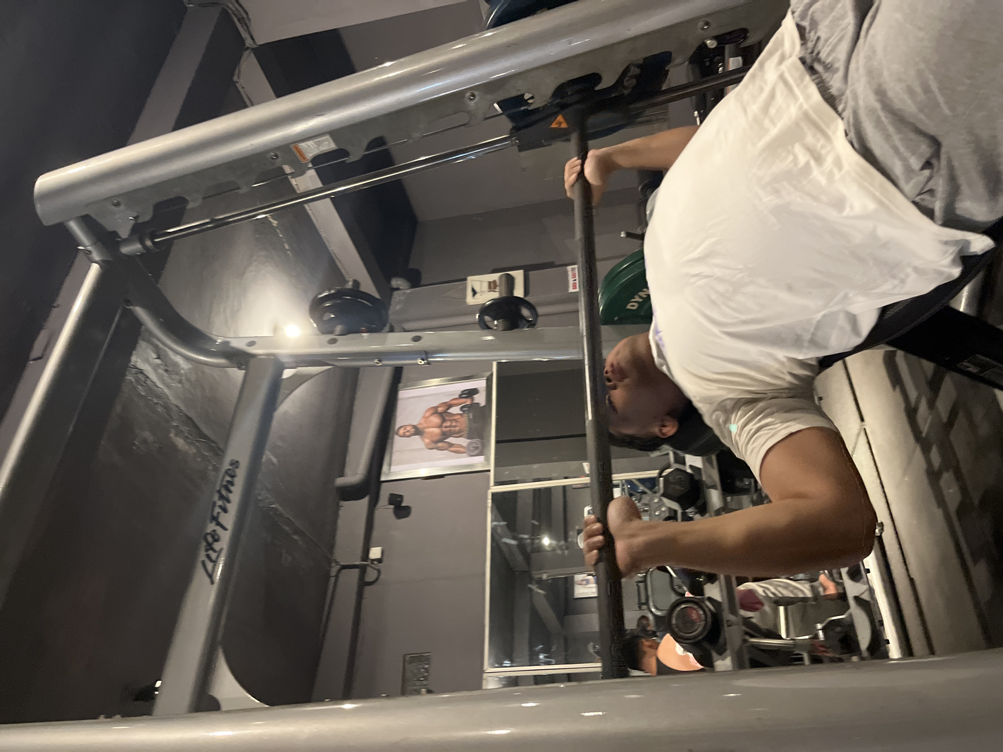 The image depicts a person exercising in a gym. The person, wearing a white t-shirt and grey pants, is positioned upside down, holding onto a horizontal bar attached to a large piece of gym equipment. The equipment is silver and appears to be a Smith machine or a similar weight training apparatus. In the background, there are other gym machines and a poster of a muscular man. The lighting suggests an indoor setting, possibly artificial gym lighting. The overall scene is one of a fitness workout. The word "LifeFitness" is visible on a piece of the gym equipment. The gym appears to be located in Denpasar, Indonesia. The image is a medium shot, focusing on the person and the gym equipment.
