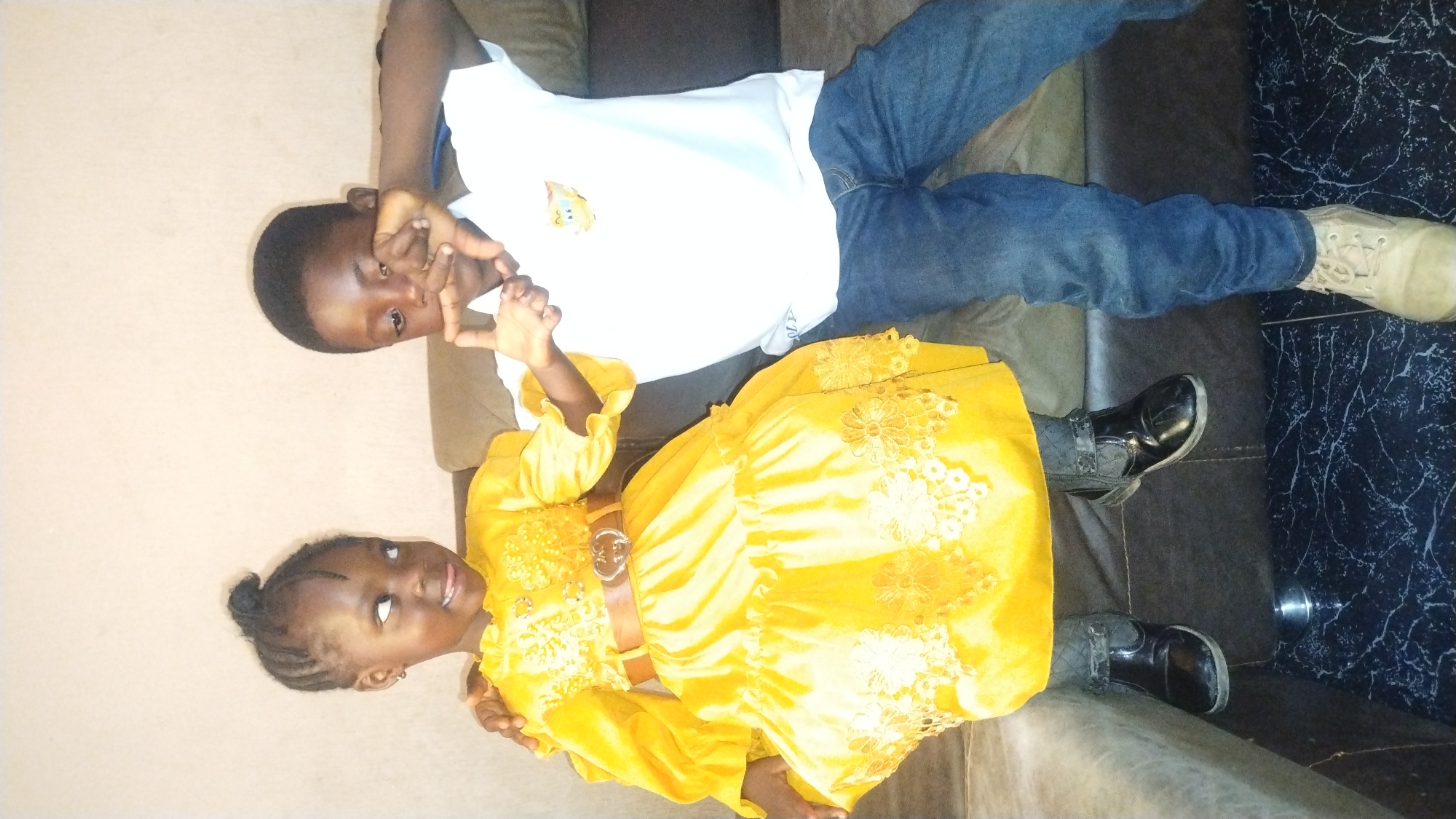 A young girl in a yellow dress and a young boy in a white t-shirt and jeans are reclining on a brown couch. The boy is making a square shape with his hands near his face. They appear to be in a room in Jos, Nigeria.