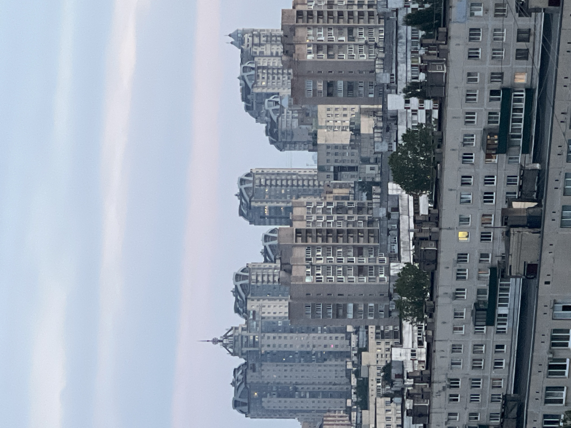 The image shows an urban skyline composed of numerous apartment buildings of varying heights and architectural styles. In the foreground, a long, low-rise apartment building with a light gray facade is prominently featured. It has multiple rows of uniformly spaced windows, some of which are illuminated, suggesting occupancy. The roofline of this building is characterized by visible ventilation shafts and chimneys.
Behind this building, a denser cluster of taller, modern apartment complexes dominates the mid-ground. These buildings feature contemporary designs with rounded or angular roof structures and numerous windows. Some of these taller buildings also show illuminated windows.
Further in the background, the silhouette of more buildings can be seen, receding into the distance. The sky above is overcast with pale blue and white clouds, indicating either dawn, dusk, or a heavily clouded day. The overall impression is a densely populated urban residential area. No people are visible in the image, and the scene is static. The location is identified as Unknown, Russia.