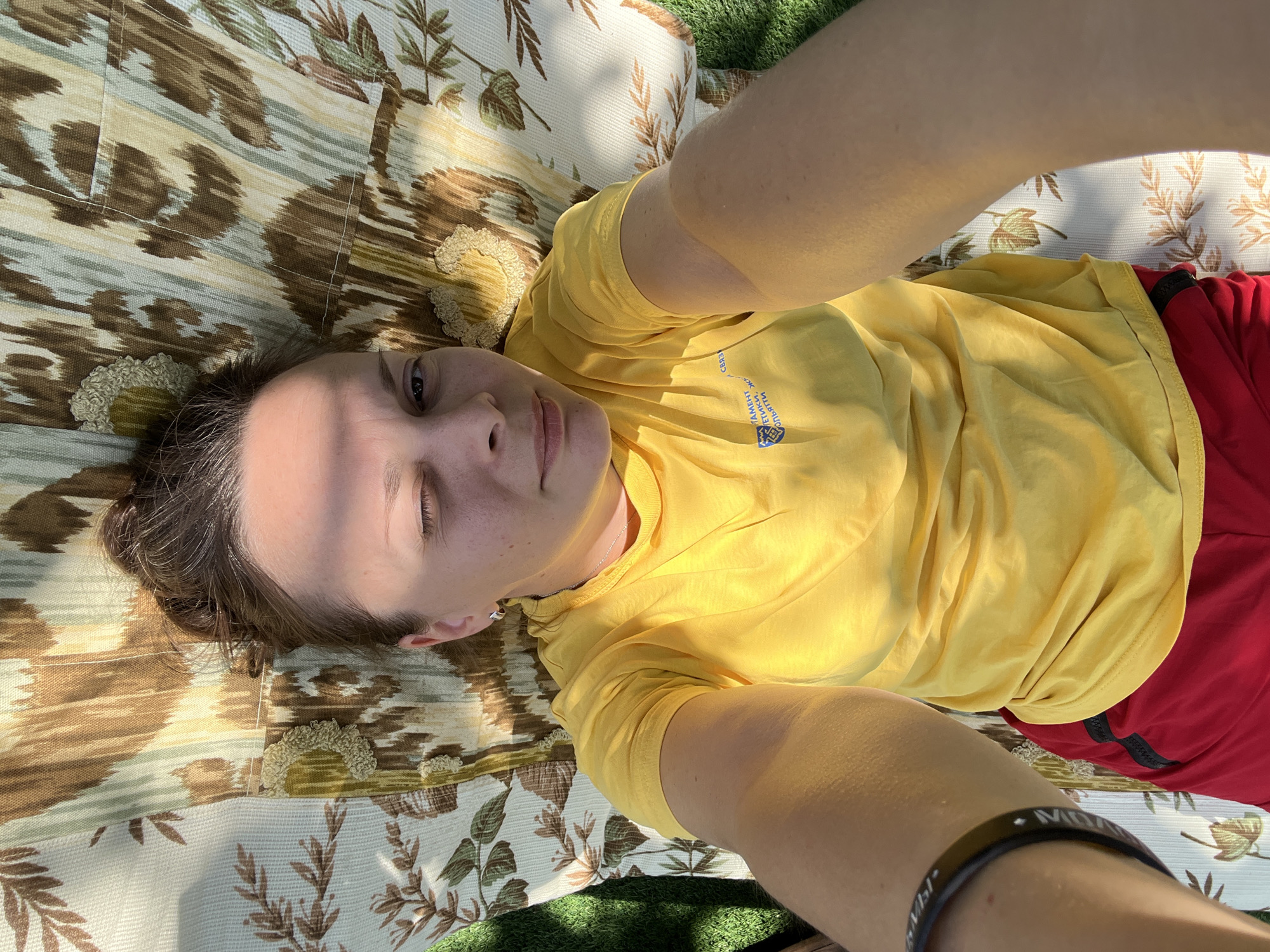 A young woman lies on her back on a floral-patterned blanket outdoors, possibly in Khryashchevka, Russia, given the sunlight and green grass visible. She is wearing a bright yellow t-shirt with a blue logo and red shorts. Her hair is pulled up in a bun. She is looking at the camera, with one eye closed and the other open, as if winking. Her arms are outstretched, holding a camera for a selfie. The lighting suggests it is a sunny day, with shadows falling across her face and arms.