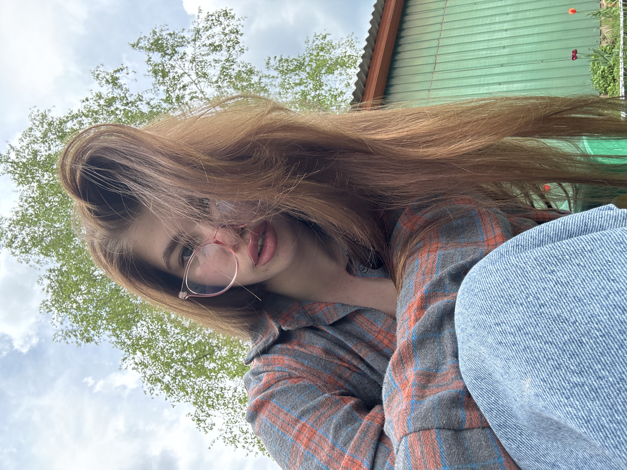 A young woman with long, red hair, wearing round pink glasses, a grey and orange plaid flannel shirt, and blue jeans, is taking a selfie from a low angle. The background shows a cloudy sky, green trees, and a green corrugated metal wall. The location appears to be outdoors, possibly in a rural or suburban area. No further specific location information related to Мещёры, Russia, is discernable.