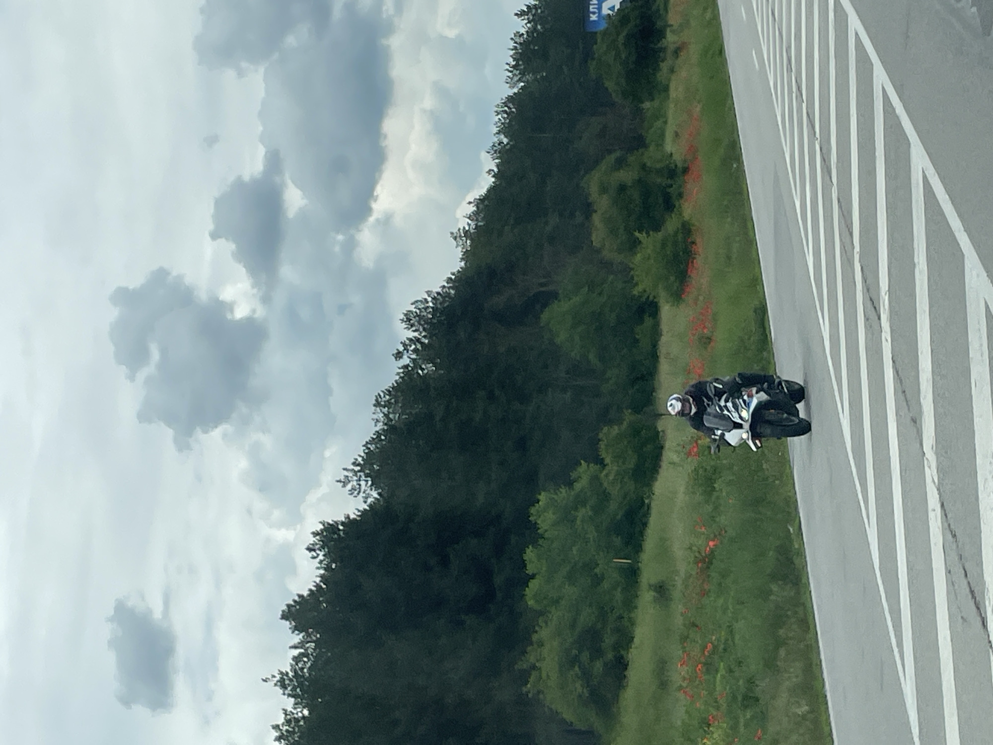 The image shows a motorcyclist on a road near Beli Lom, Bulgaria. The rider, wearing a helmet and dark clothing, is on a light-colored motorcycle. The road has white lane markers. To the left of the road is a grassy area with red flowers and trees. A forest is in the background. The sky is cloudy.