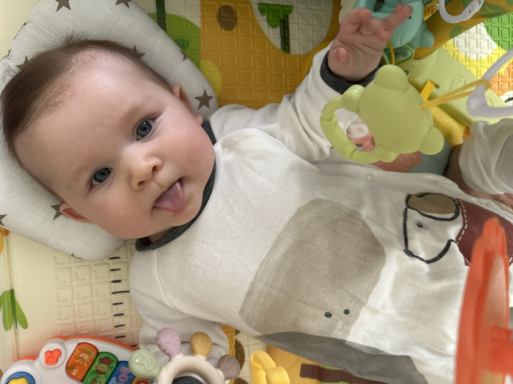 A baby lies on a padded playmat, resting on a pillow with star patterns. The baby is wearing a light-colored onesie with a brown elephant design. Toys surround the baby, including a hanging mobile with a green frog, a keyboard, and various rattle toys. The location appears to be indoors, possibly a home in Chaikovsky, Russia, although there is no definitive information to confirm the city.
