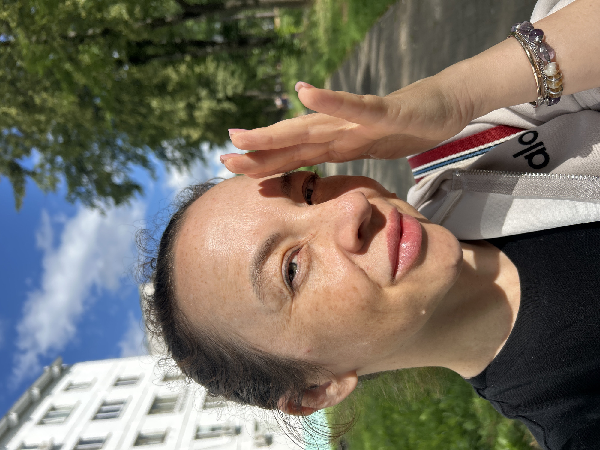 A close-up, portrait orientation photograph shows a woman shielding her eyes with her hand from a bright sun. She is wearing a white zip-up hoodie with red and blue trim, and a black t-shirt underneath. A multi-colored beaded bracelet adorns her left wrist. Her skin has a light dusting of freckles. In the background, a white building with multiple windows is visible, along with lush green trees and a bright blue sky with scattered white clouds, suggesting a sunny day. The overall impression is one of a casual outdoor moment. The text "alo" is visible on the hoodie. The photo appears to be taken during the daytime.