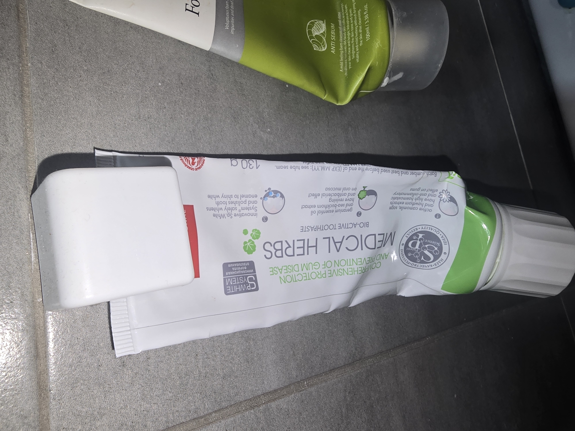 The image shows two tubes of toothpaste standing upright. The tube on the left is white with green and red text, and has a white cap resting on its side near the top. The text on this tube indicates it is "MEDICAL HERBS BIO-ACTIVE TOOTHPASTE" and features a "WHITE SYSTEM". The tube on the right is green and white, with text indicating it is a "SEBUM" product for the face. The background appears to be a tiled wall. This scene is not identifiable as being in Ulan-Ude, Russia.