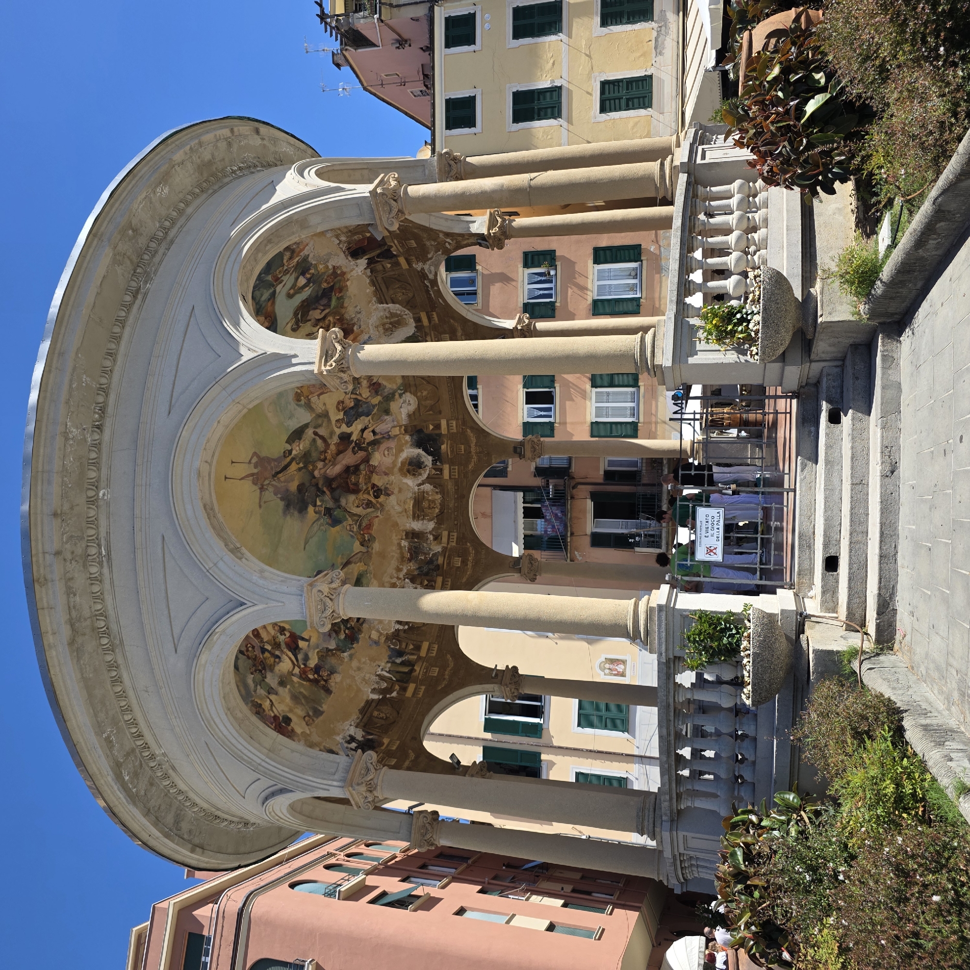 The image shows the iconic Loggia of Victory in Rapallo, Italy, a historic circular monument featuring a frescoed ceiling and supported by columns. The loggia is situated in a public area with surrounding buildings and lush greenery. Several individuals are visible, some standing near the entrance of the loggia, while others are in the background, suggesting a lively atmosphere. The fresco depicts a scene with multiple figures in dynamic poses, possibly representing a historical or mythological event. The architecture of the loggia, with its ornate detailing and classical columns, stands out against the backdrop of colorful buildings, characteristic of Italian urban landscapes. The scene is bathed in bright sunlight, casting distinct shadows and highlighting the vibrant colors of the surroundings.