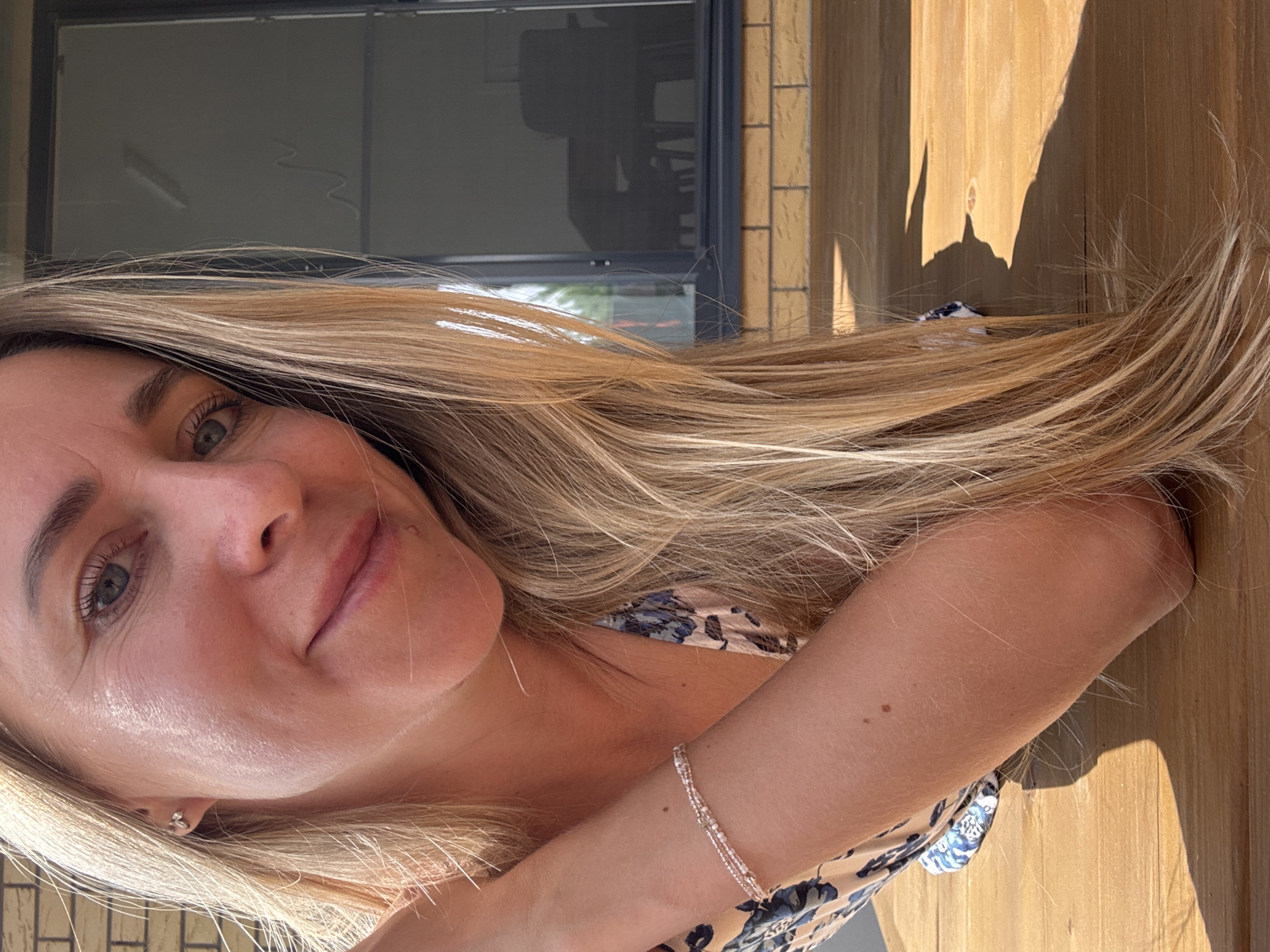 A woman with long blonde hair smiles at the camera in a close-up shot. She is wearing a floral print top and a beaded bracelet on her left wrist. Her right arm is bent, resting on a wooden surface. The background shows a brick wall and a glass door. The scene appears to be outdoors, possibly on a deck or patio. The city of Chaikovsky, Russia, is not depicted in this image.