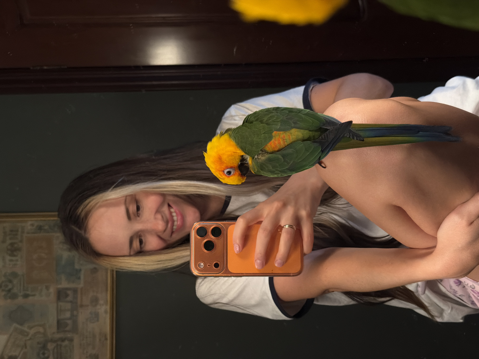A young woman with blonde-streaked brown hair smiles as she holds up an orange smartphone to take a selfie. A green and yellow parrot is perched on her arm, its head tilted as if looking at the camera. The woman is wearing a white t-shirt with dark trim. The scene appears to be indoors, with dark wood paneling visible in the background, possibly a mirror or furniture. There is also a framed picture on the wall to the left. The overall mood is casual and playful, suggested by the woman's smile and the presence of the bird. The lighting is soft, and it's difficult to determine the time of day. The image is oriented vertically, with the subjects facing the right side of the frame.