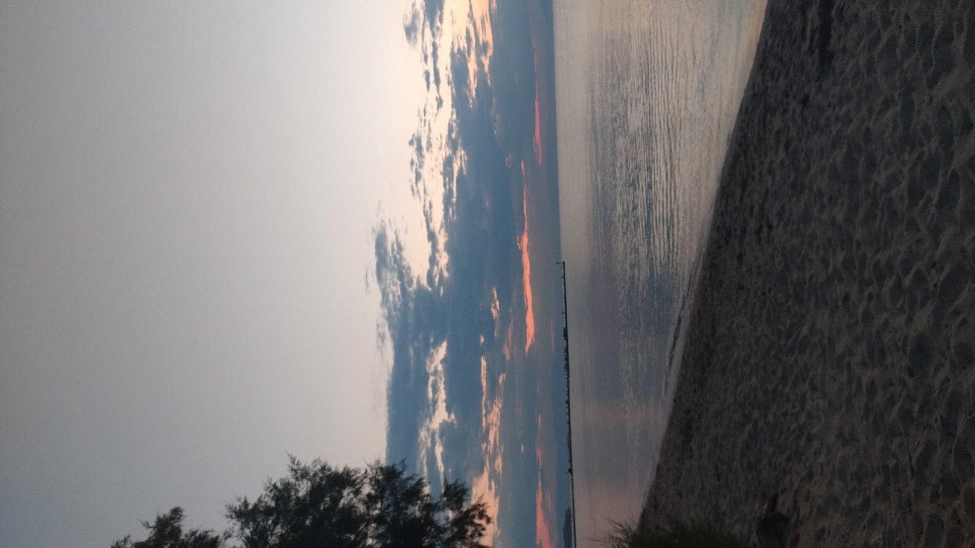 A vertical, low-angle, full shot captures the twilight beach scene in Neoi Epivates, Greece. The sky transitions from a pale grey at the top to a warmer, soft orange hue near the horizon, punctuated by scattered, dark grey clouds. A thin, orange band of light is visible just above the horizon. The sea is calm, with subtle ripples reflecting the muted sky colors. On the left side of the frame, the dark silhouette of a tree with sparse foliage extends from the top down. The foreground is a sandy beach, its texture and ripples from foot traffic or wind clearly visible in the dim light. In the distance, a long pier or breakwater stretches across the water, lined with small structures or lights and culminating in a visible lighthouse. The overall atmosphere is serene and quiet, typical of a beach at dusk. No people are present in the image.