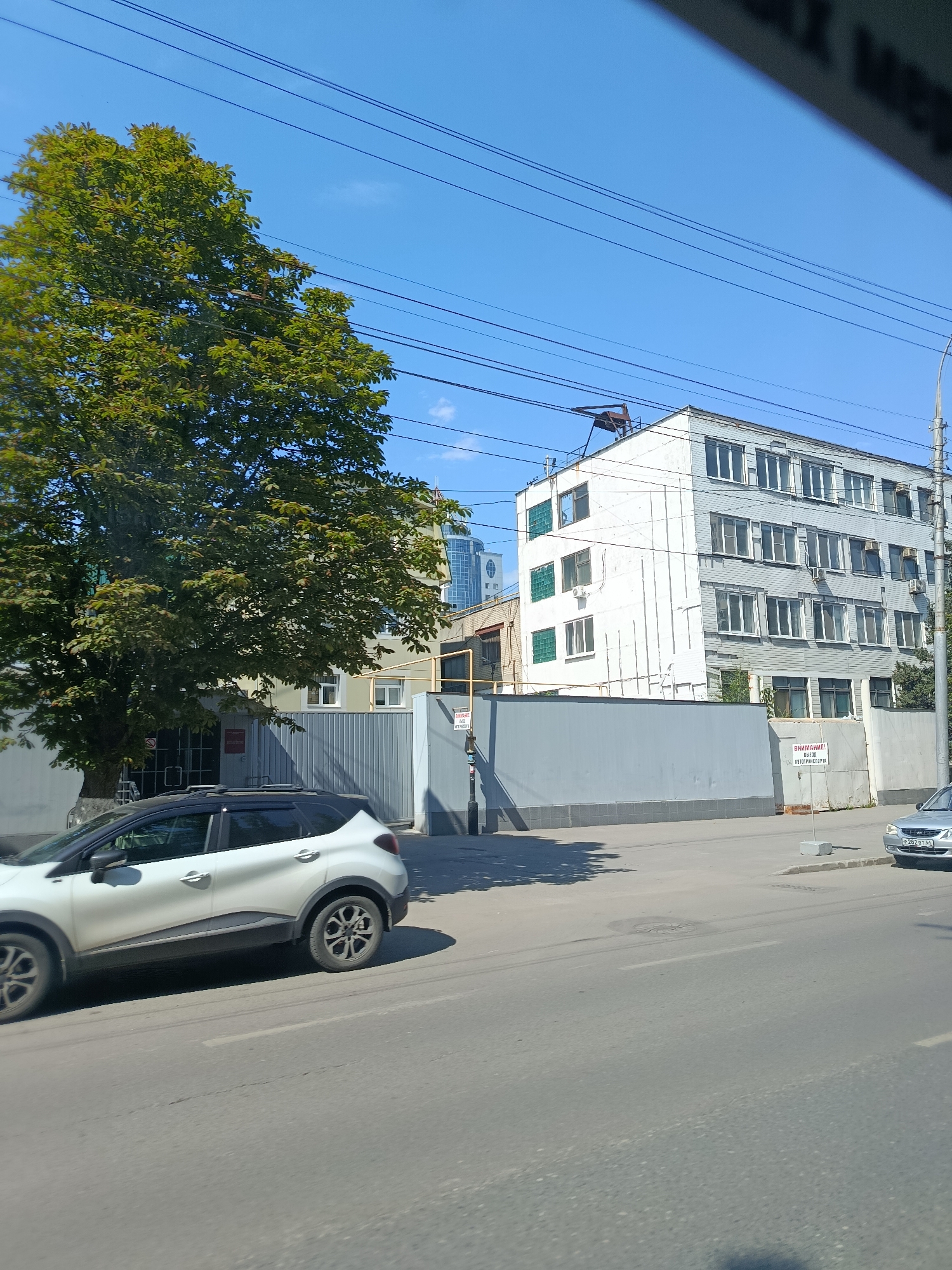 The image depicts a view from a moving vehicle on a street in Saratov, Russia. In the foreground, a white Renault Captur SUV is partially visible, positioned on the right side of the road. The street is paved with asphalt, and visible road markings include a dashed white line.
To the left of the street, a large tree with green foliage stands prominently. Behind the tree, a grey metal fence encloses a property. A multi-story building with a light-colored facade and numerous windows is visible beyond the fence. This building appears to be an industrial or office building. Further in the background, a modern skyscraper with a glass facade can be seen.
Above the buildings and tree, a clear blue sky is visible, with several overhead power lines stretching across the frame. A smaller, silver car with a license plate is parked on the right side of the road, near a concrete barrier. A warning sign with Russian text, "ВНИМАНИЕ! ВЫЕЗД" (ATTENTION! EXIT), is affixed to a concrete post near the fence. The overall scene suggests an urban environment with a mix of older and newer architecture, under a bright, sunny sky.