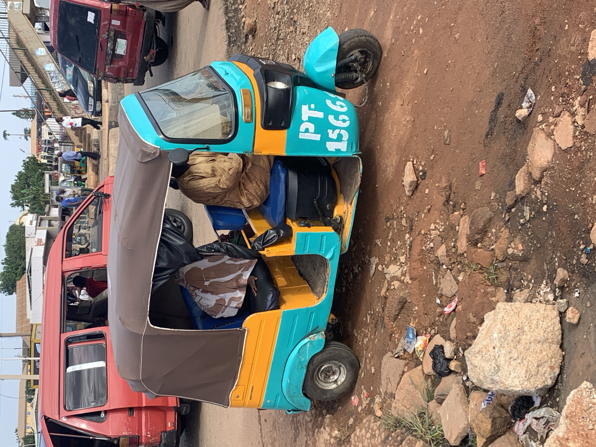 The image captures a scene in the city of Gura Top, Nigeria, focusing on a three-wheeled auto-rickshaw, commonly known as a "Keke." The Keke is painted in vibrant colors, with a turquoise lower body and a yellow stripe on the upper part, topped by a brown canopy. It is parked on a dirt and rocky road, with a red minivan visible behind it. A person is seated in the driver's position of the Keke, wearing a brown robe-like garment. Another person is partially visible in the passenger area, with a patterned cloth draped over them. The Keke displays "PT-1566" in white lettering on its side. The surrounding environment shows a dusty, unpaved road with scattered rocks and debris, suggesting an outdoor, possibly informal, area. In the background, various vehicles, buildings, and people are present, indicating a busy urban setting. The overall impression is of a typical street scene in a Nigerian city, with the Keke as the central element.