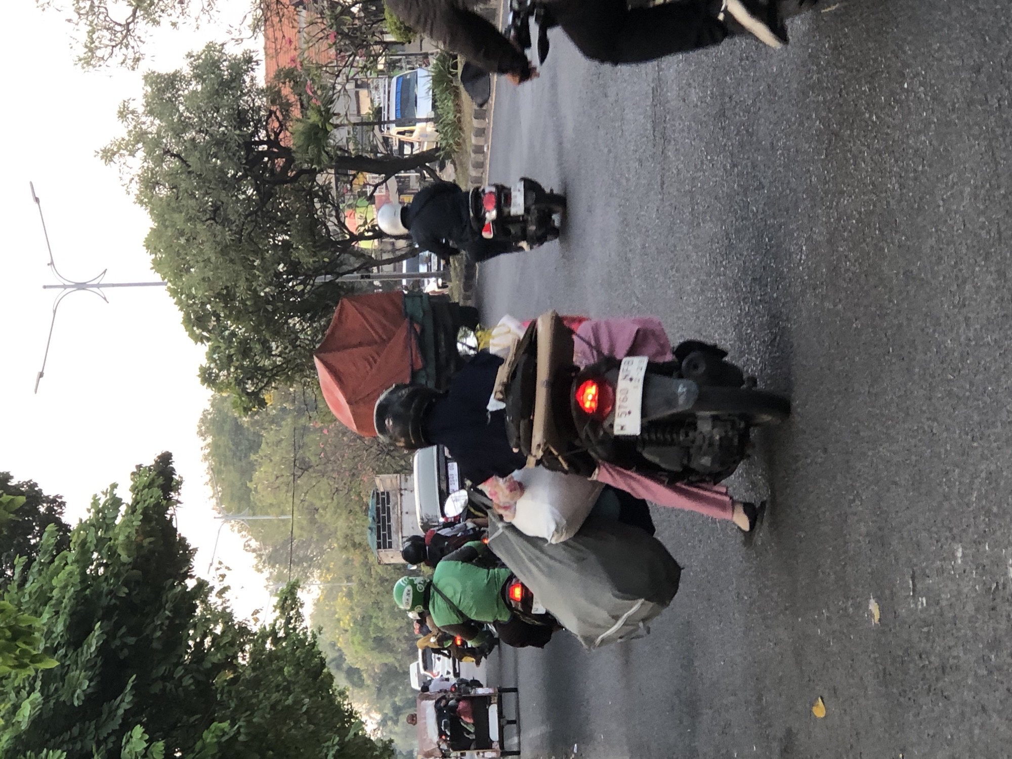 In Sumorame, Indonesia, a busy street scene unfolds with several motorbikes navigating traffic. In the foreground, a person wearing pink pants and a pink cloth is riding on the back of a motorbike, carrying a large gray bag and a white bag. The motorbike's license plate reads "N 5760 BB". To the left, another motorbike with a sidecar is visible, carrying luggage. Further ahead, a motorbike with a green jacket and helmet, possibly a delivery driver, is also in motion. In the background, trucks and other vehicles are seen on the road, with trees and buildings lining the street. The overall atmosphere suggests a typical day with considerable traffic.