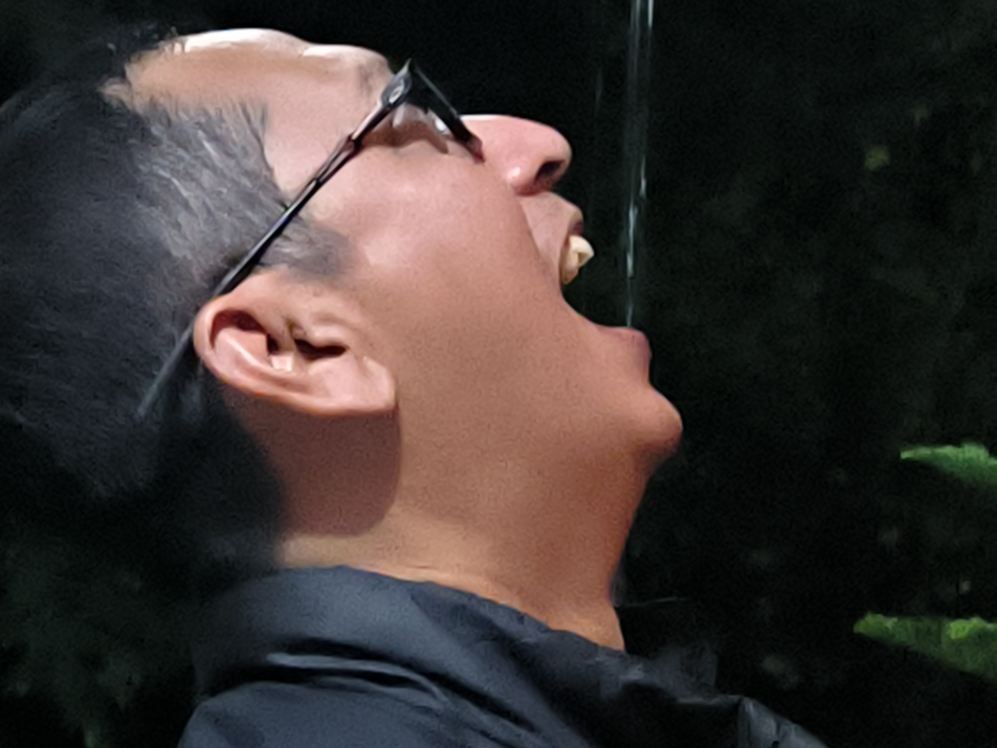A close-up shot reveals a man with dark hair, glasses, and brown skin, drinking water from a source above him. His head is tilted back, mouth open. The background is dark with green foliage, potentially indicating an outdoor setting. The location in Lhokseumawe, Indonesia, is not evident from the image.
