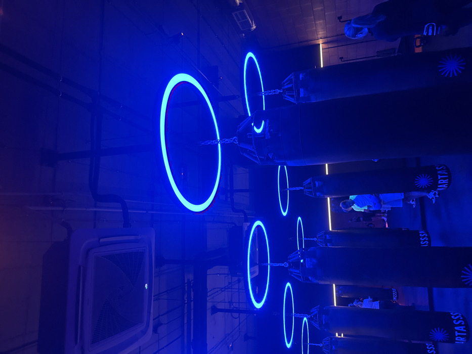 A row of boxing bags hangs in a dimly lit room with blue neon rings. The bags are suspended by chains. Text is visible on the bags. People are present in the background. An air conditioning unit is visible on a wall with pipes running along the wall. The rings appear to be spaced equally in front of each boxing bag, creating a visually rhythmic pattern.