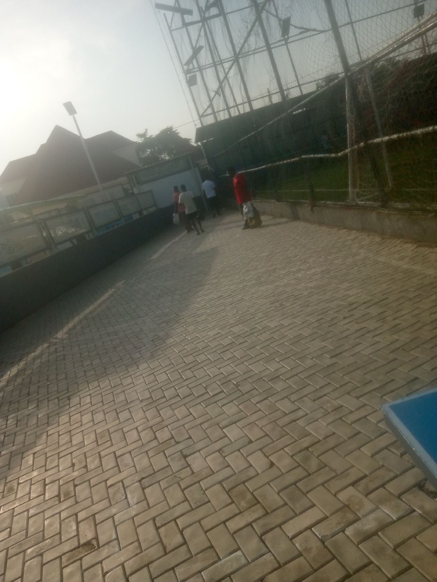 The image captures a sunlit outdoor scene on a paved walkway. The walkway is constructed with rectangular tan pavers arranged in a herringbone pattern. In the background, there are several people walking. One individual in the foreground is wearing a red shirt and carrying a bag. To the left of the walkway, a dark grey wall lines the area, and behind that, a building with a reddish-brown roof is visible. A tall lamppost stands near the building. Further back, a large net-covered structure, possibly a sports court, dominates the upper portion of the frame, with what appears to be a grass field visible beneath it. The sky is bright and hazy, suggesting it is either morning or late afternoon. The overall impression is a public area, possibly a park or recreational facility, in the city of Jalingo, Nigeria.