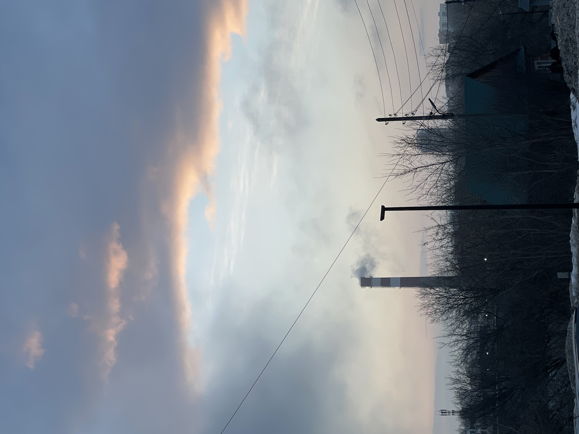 The image depicts an overcast sky in Izhevsk, Russia, during what appears to be either sunrise or sunset. The sky is filled with various shades of gray, with some pink hues on the clouds. In the foreground, bare trees and utility poles with overhead wires are visible. A tall industrial chimney with a red and white pattern emits smoke. A residential building can be seen in the distance. The photograph has a slightly tilted perspective.