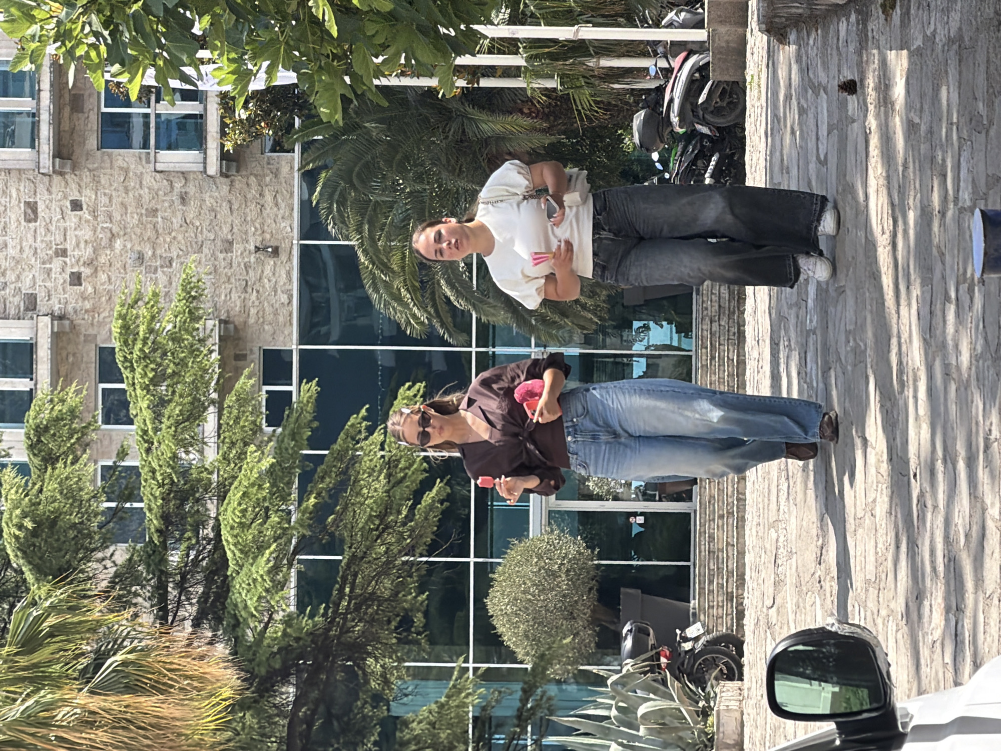 The image shows two young women standing outside a building. The woman on the right is wearing a white t-shirt and jeans, and is holding a pink fan. The woman on the left is wearing a brown top and jeans, and is also holding a pink fan. They are both looking at the camera and smiling. The scene appears to be set in a sunny location, possibly a tourist area, with palm trees and a building with stone cladding. There is a motorcycle parked in the background, and a car mirror is visible in the foreground. The image is taken vertically and the subjects are slightly blurred.