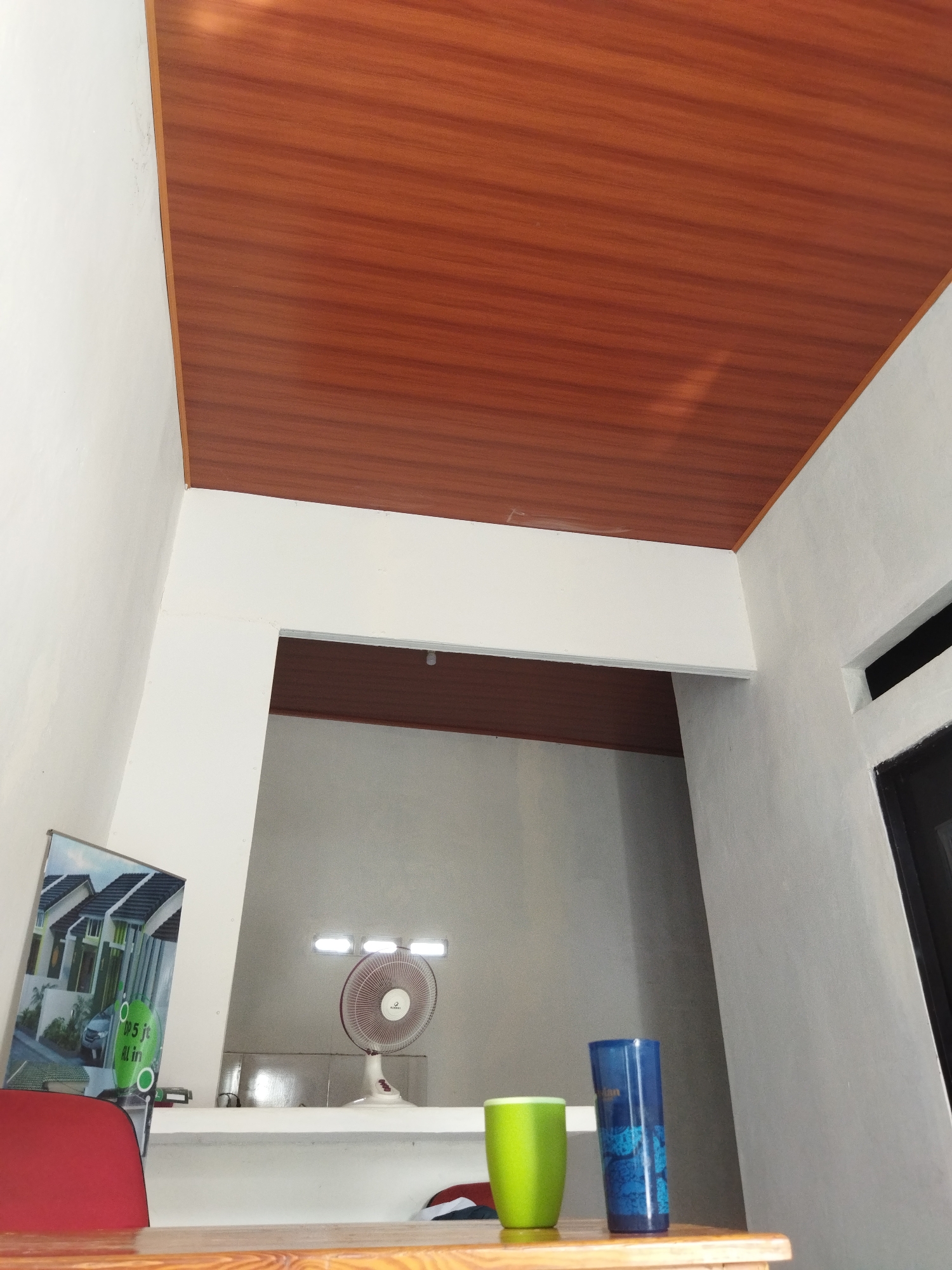 The image shows the interior of a room with white walls and a wood-paneled ceiling. In the foreground, there is a wooden table. On the table, there are two cups, one green and one blue. Behind the table, there is a white shelf. On the shelf, there is a white fan with a red trim and a picture of houses. There are also some lights on the wall. The image appears to be taken in Suka Sari, Indonesia.